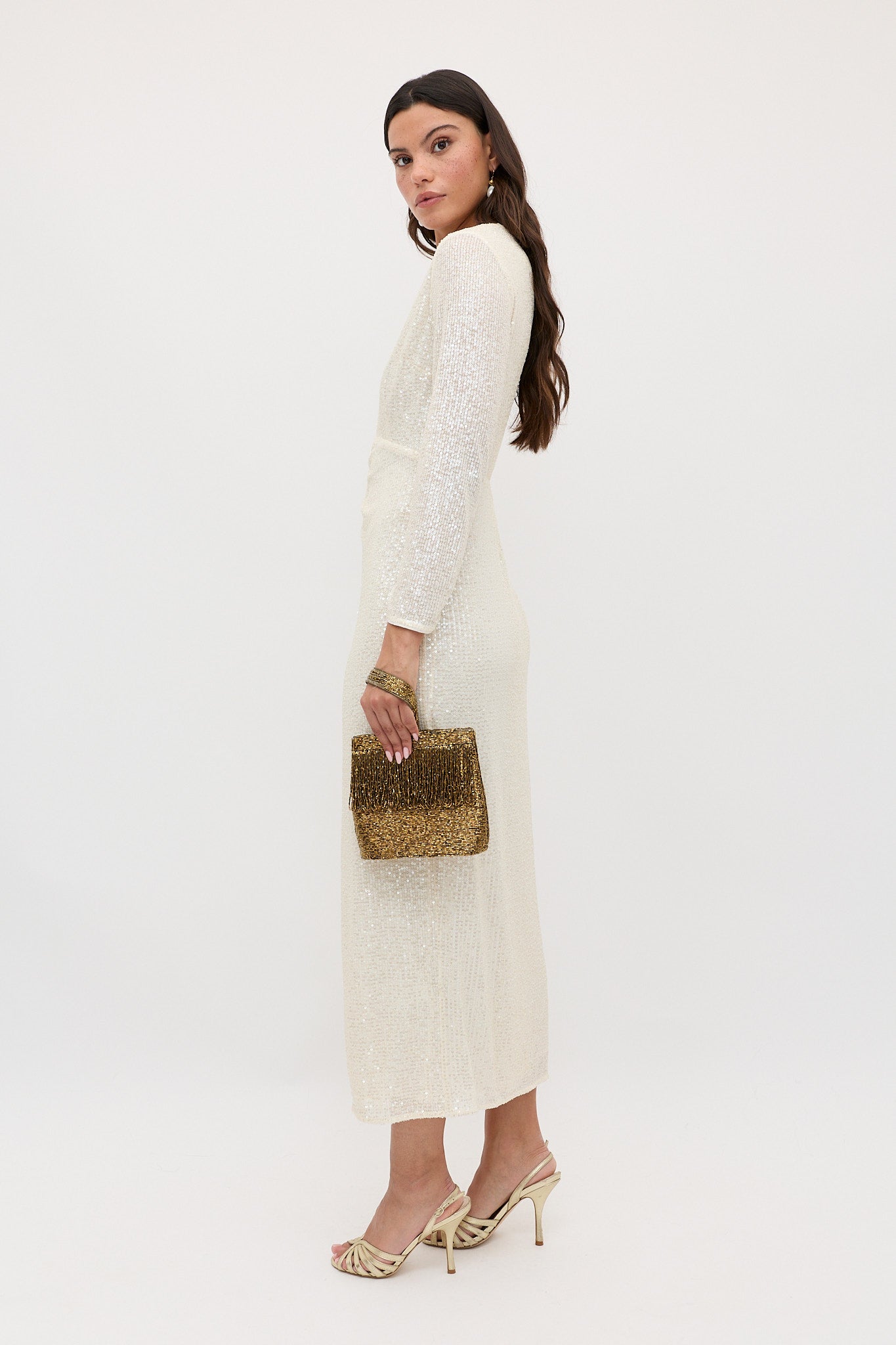 A woman stands sideways in the Lorenzo - Pearl long-sleeved, shimmery ivory dress, holding a gold beaded clutch. She wears heeled sandals, has long wavy brown hair, and poses against a plain white background.