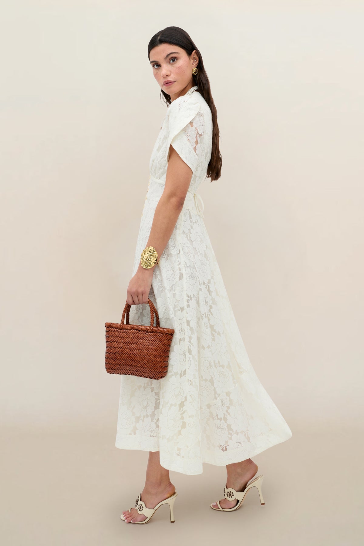 A woman in the Novara - Ivory white floral lace midi dress with curved sleeves stands sideways, accessorized with gold earrings, a large gold bracelet, a small woven brown handbag, and open-toe heeled sandals against a plain background.