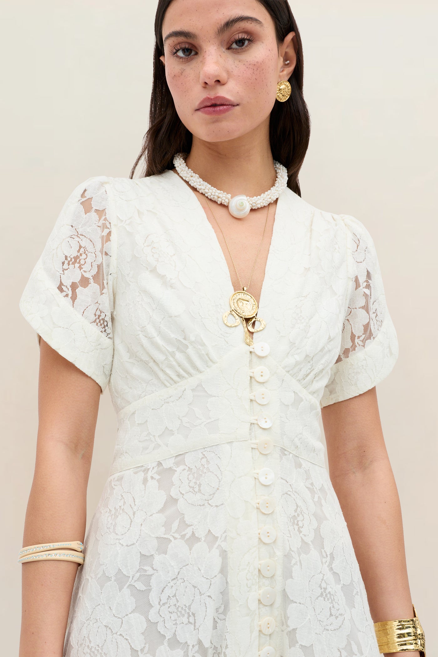 A woman in the Novara - Ivory vintage floral lace dress with curved short sleeves and front buttons, accessorized with layered gold necklaces, a pearl choker, hoop earrings, and gold bangles, stands against a neutral background.