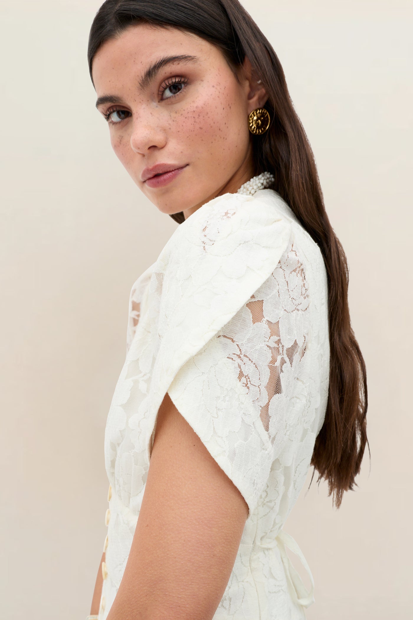 A woman with long brown hair and freckles wears the Novara - Ivory vintage dress with floral lace, gold earrings, and a pearl necklace, gazing over her shoulder against a plain light background.