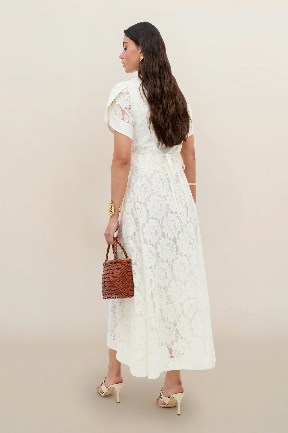 A woman with long brown hair faces away, wearing the Novara - Ivory floral lace short-sleeve dress, heeled sandals, gold jewelry, and holding a small brown woven handbag against a plain beige background.
