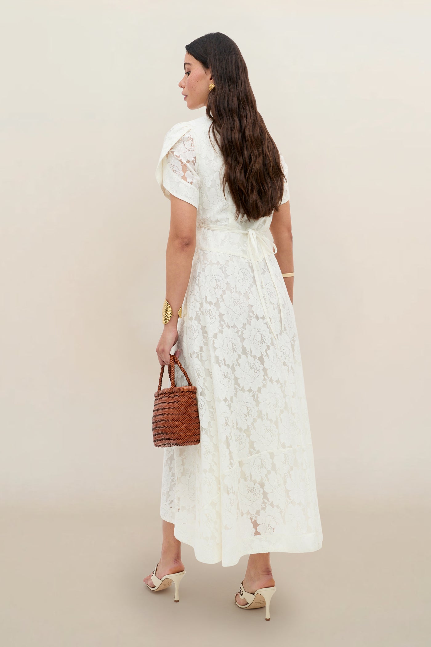 A woman with long brown hair faces away, wearing the Novara - Ivory floral lace short-sleeve dress, heeled sandals, gold jewelry, and holding a small brown woven handbag against a plain beige background.