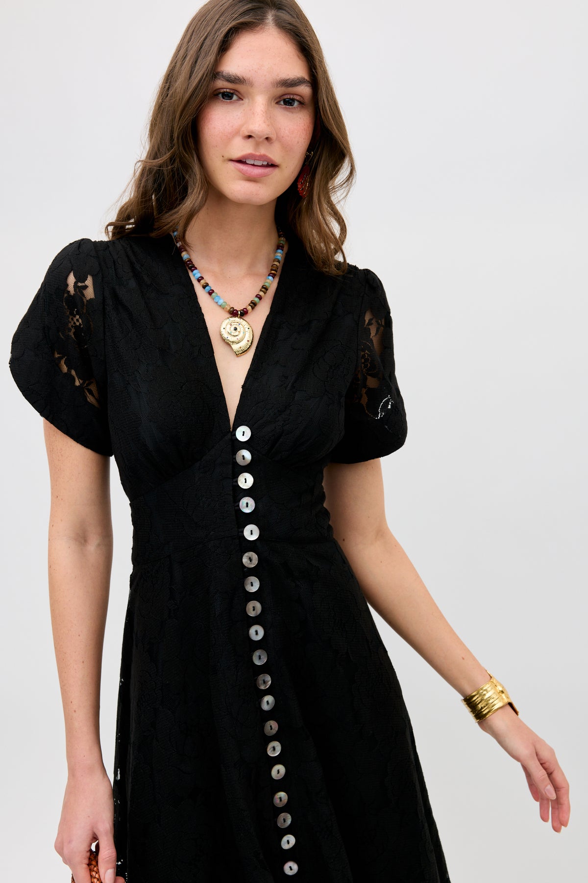 A woman wearing the Novara - Black dress with short sleeves and large front buttons stands against a plain background. She has long brown hair, a colorful beaded necklace, and a gold bracelet.