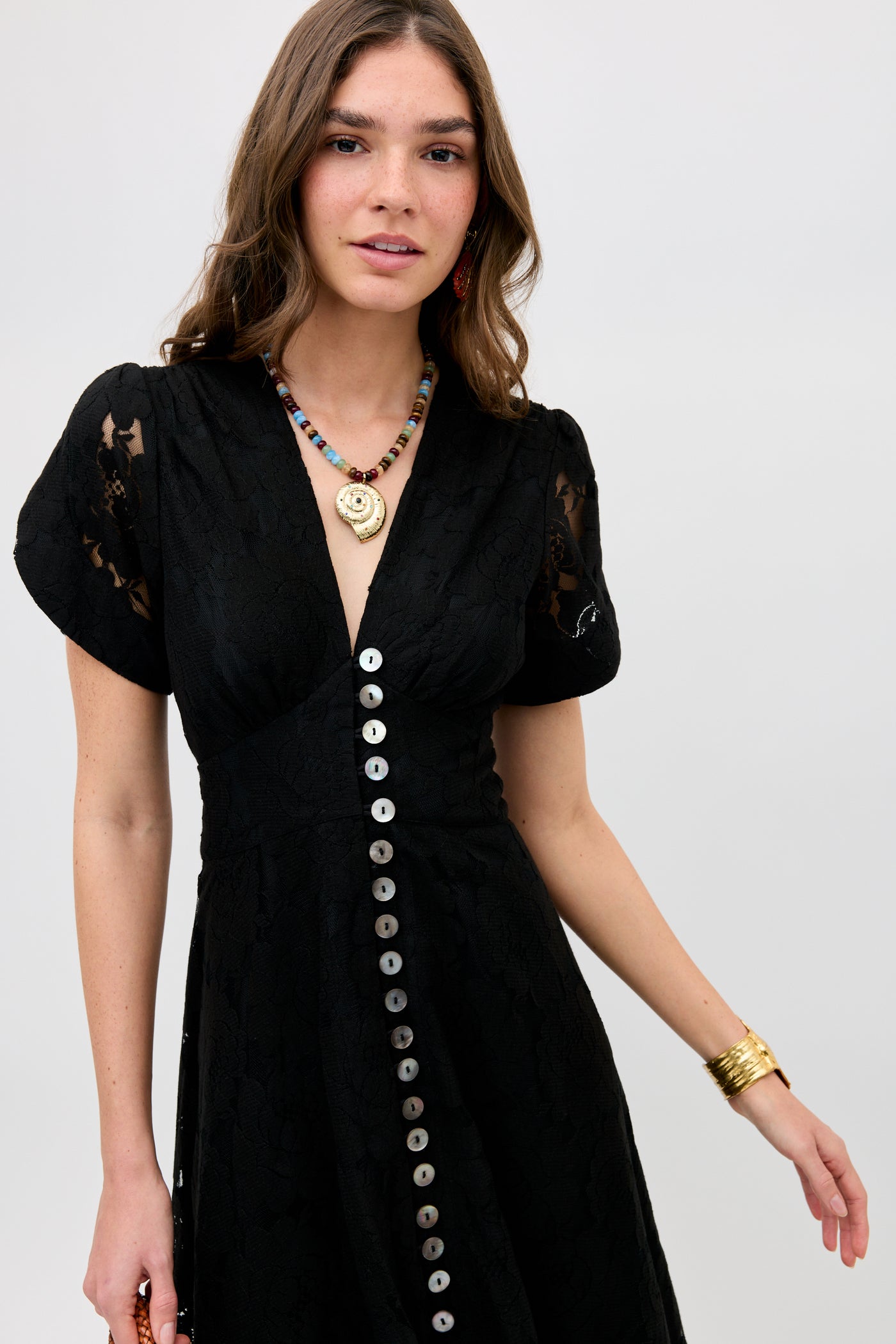 A woman wearing the Novara - Black dress with short sleeves and large front buttons stands against a plain background. She has long brown hair, a colorful beaded necklace, and a gold bracelet.