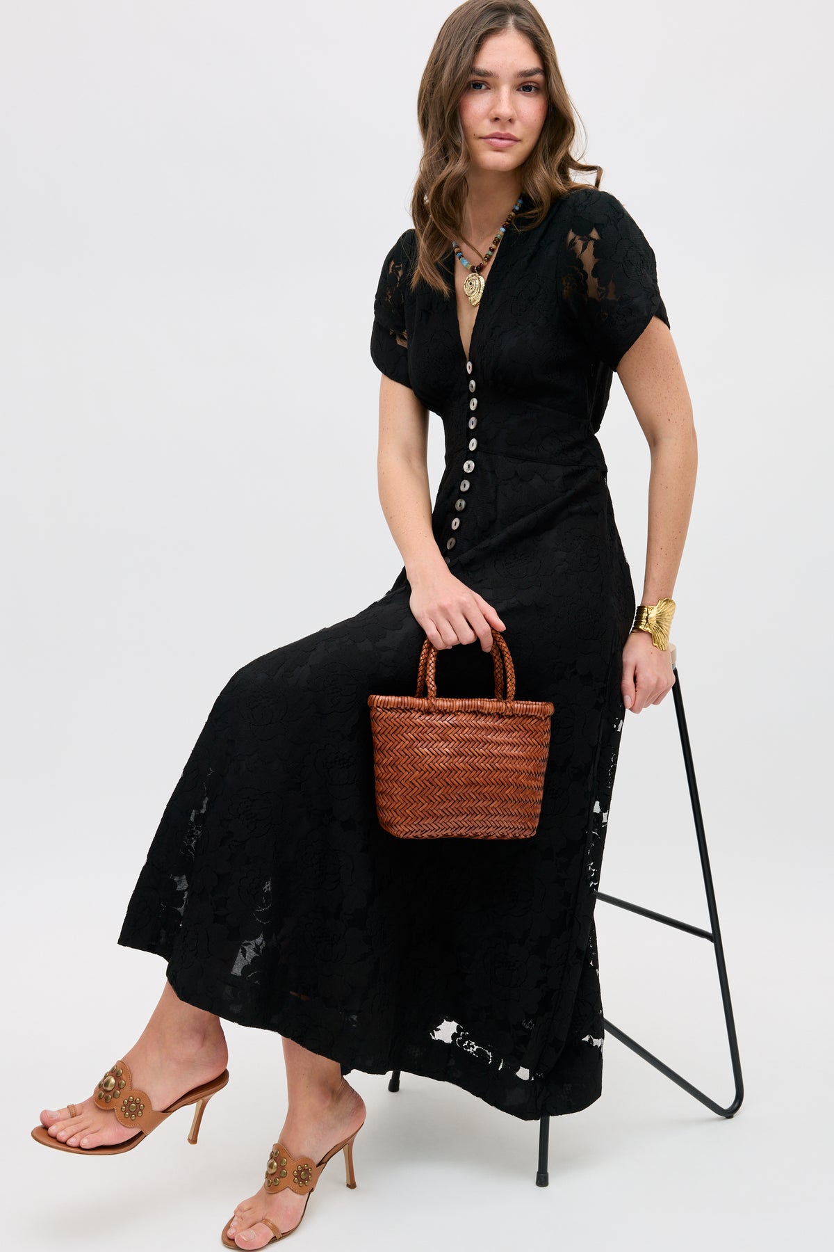 A woman wears the Novara - Black button-down lace dress, seated on a black stool with a woven brown handbag. She pairs it with tan heeled sandals, gold jewelry, and loose brown hair against a plain, light background.