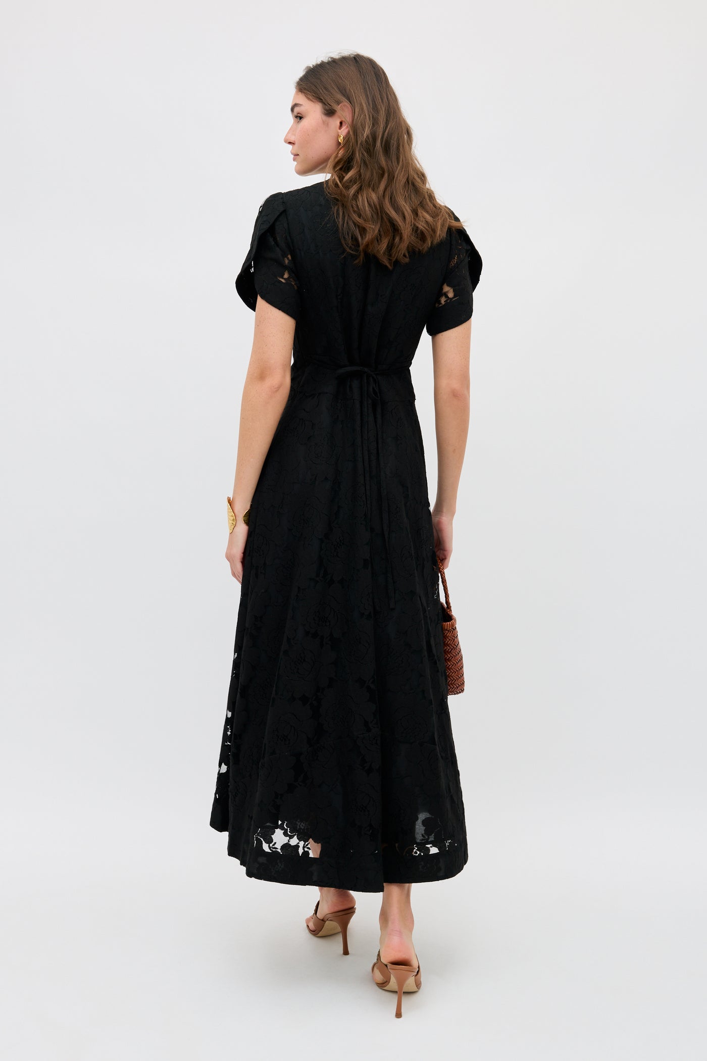 A woman with loose, wavy brown hair faces away, wearing the Novara - Black lace midi dress with short sleeves and a waist tie. She carries a brown woven handbag and pairs the look with tan heeled sandals.