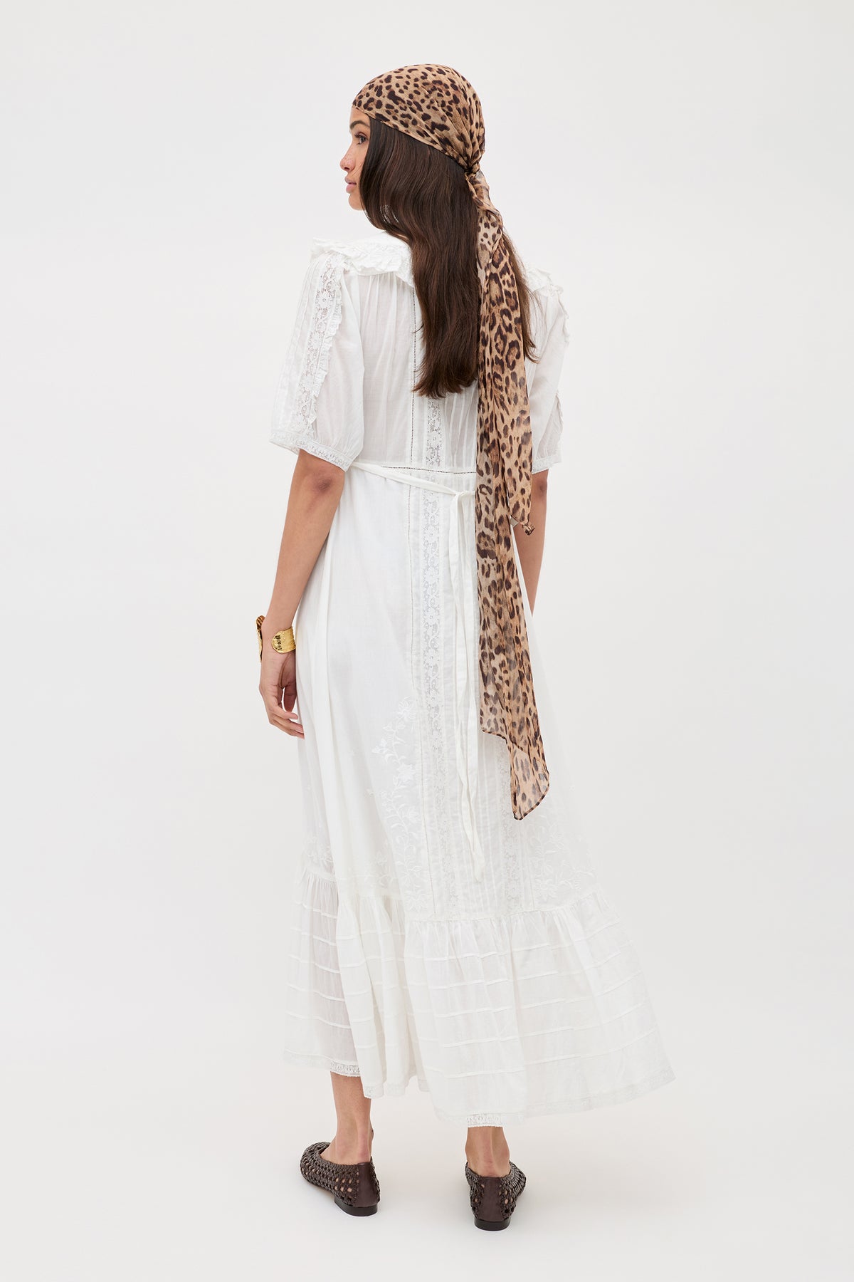 A woman stands facing away in the Edina - Pansy Embroidery White dress, with delicate lace embroidery, paired with brown flats, a gold bracelet, and a leopard-print headscarf, set against a plain white background.