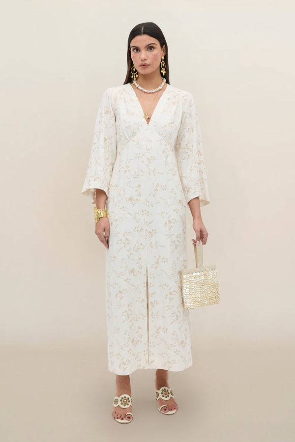 A woman stands against a plain background wearing the “Dahlia - Botanical Embroidery Pearl” dress, featuring vintage-inspired floral embroidery and flared sleeves, styled with gold jewelry, white heeled sandals, and a woven square handbag.