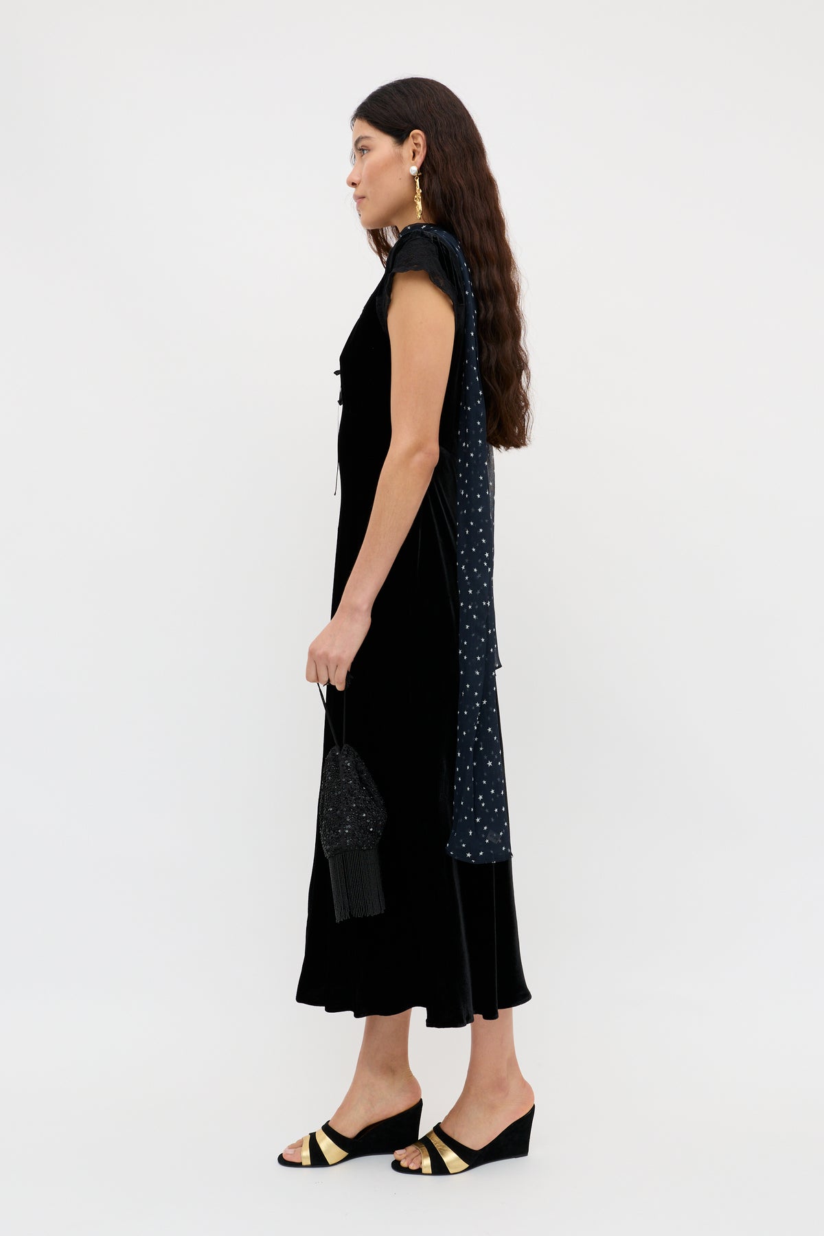 A woman with long brown hair stands in profile against a white background, wearing the Amari - Black sleeveless velvet dress, a star-patterned scarf, black and gold sandals, and holding a small black purse.