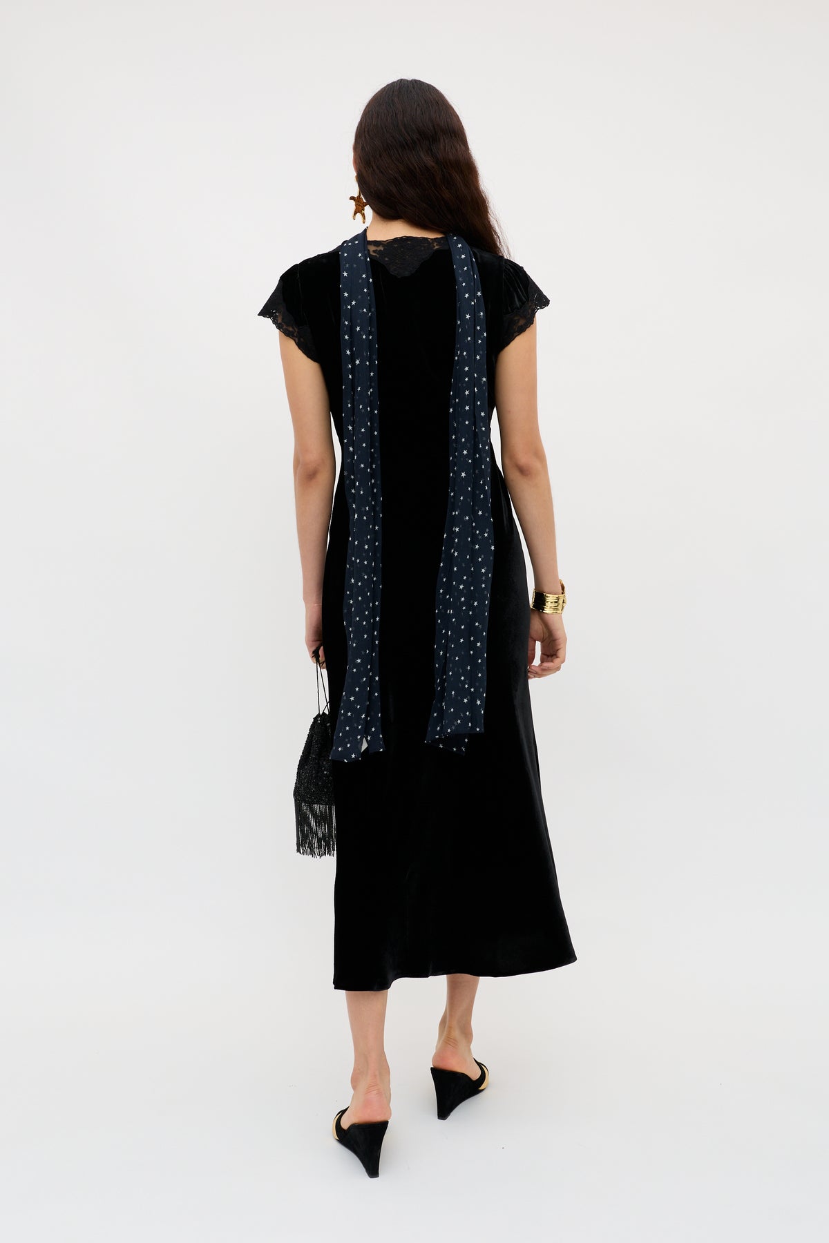 A woman with loose hair wears the Amari - Black velvet dress with lace sleeves, a long blue patterned scarf, black pointed heels, a black fringed handbag, and a gold watch.