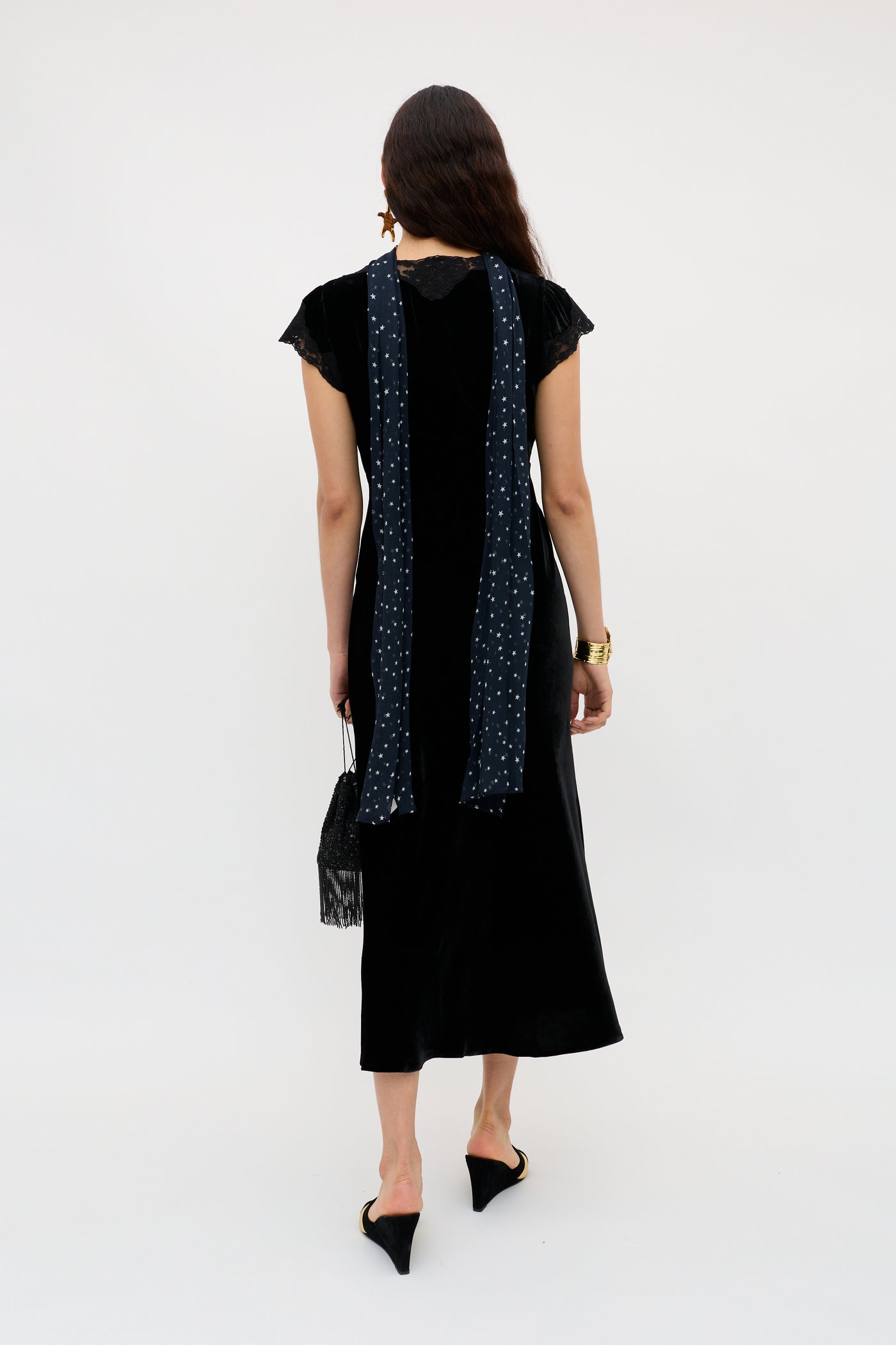 A woman with loose hair wears the Amari - Black velvet dress with lace sleeves, a long blue patterned scarf, black pointed heels, a black fringed handbag, and a gold watch.