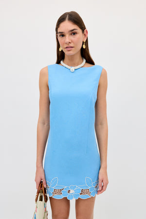 A woman wears the Bingley - Daisy Cutwork Embroidery Blue sleeveless linen dress, paired with a beaded necklace and gold earrings, standing against a white background while holding a white bag.