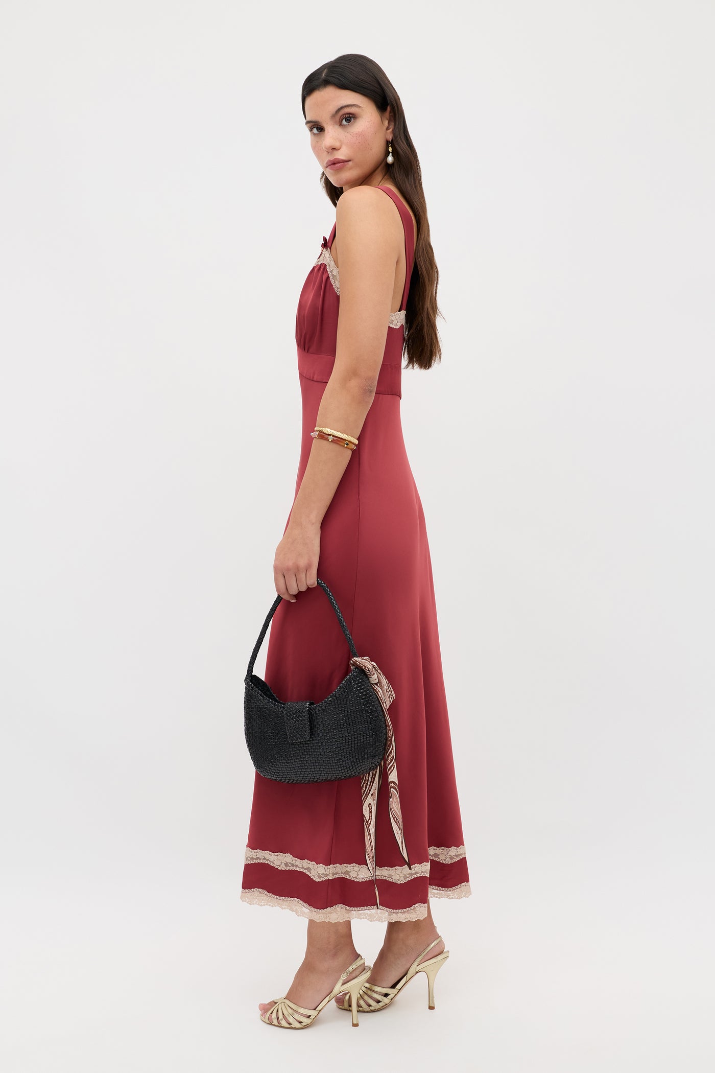 Wearing the Lucie - Rose, a sleeveless ankle-length maroon dress with lace trim, a woman stands sideways with a black textured handbag. She pairs gold heels, a gold bracelet and earrings, her long dark hair styled loosely.