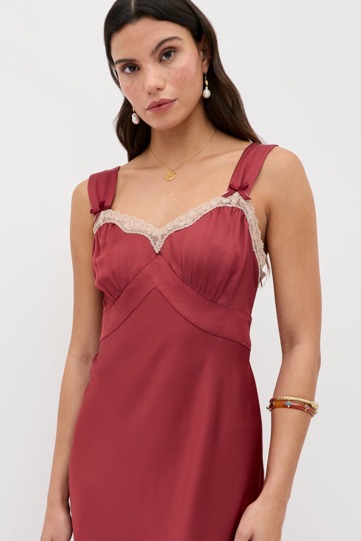 A woman wearing the Lucie - Rose sleeveless dress, featuring lace trim and tied shoulder straps. She has long brown hair, gold jewelry, and a neutral expression, standing against a plain light background.