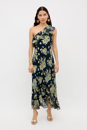 A woman stands against a plain white background wearing the Leon - Bouquet Devoré Navy, a navy blue one-shoulder ruffled midi dress with yellow and green floral print, paired with gold earrings, a gold bracelet, and open-toe heels.