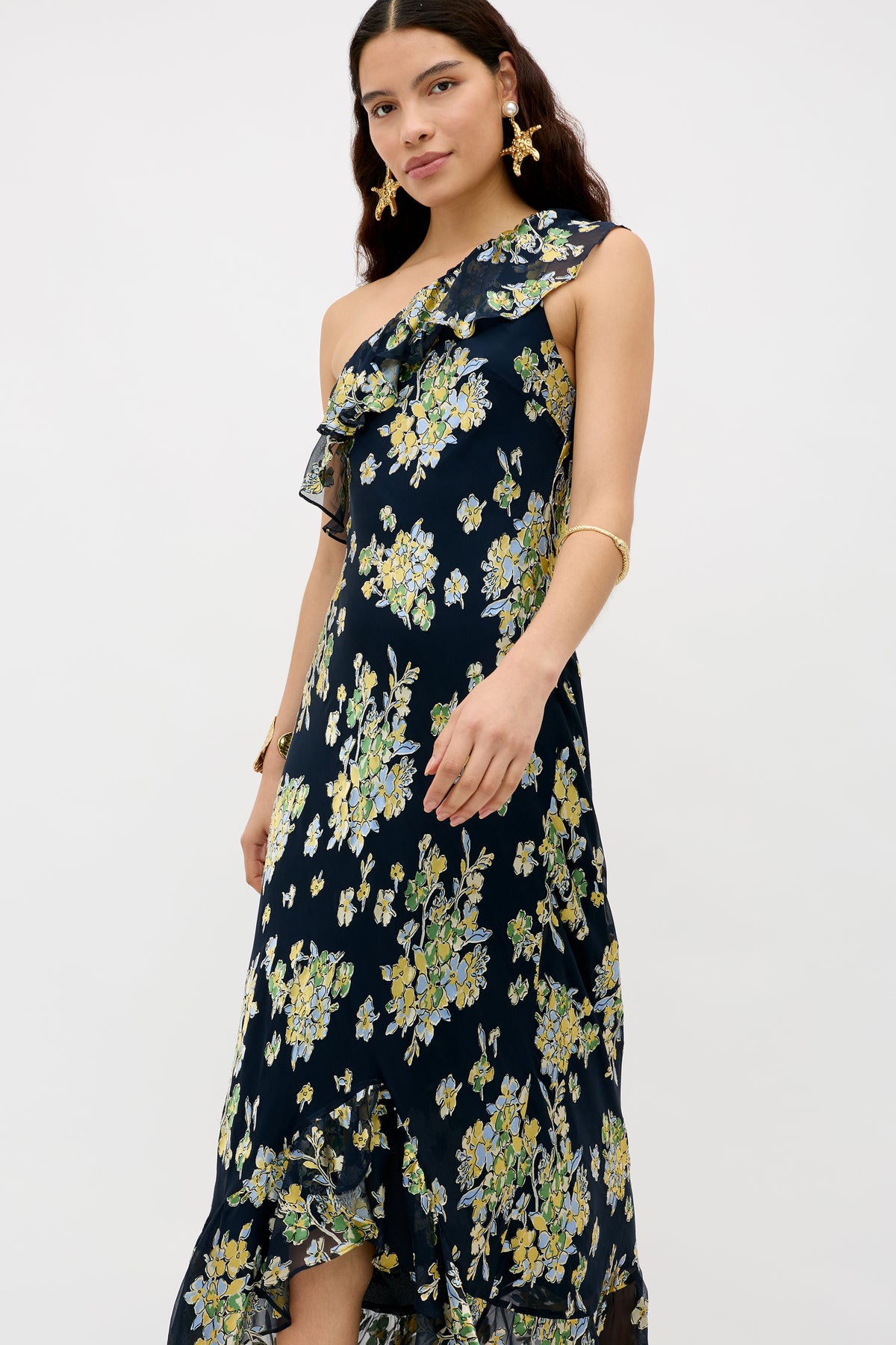 Woman wearing the Leon - Bouquet Devoré Navy one-shoulder dress with yellow and green floral print and ruffled neckline, paired with gold earrings and bracelet, standing against a plain white background.