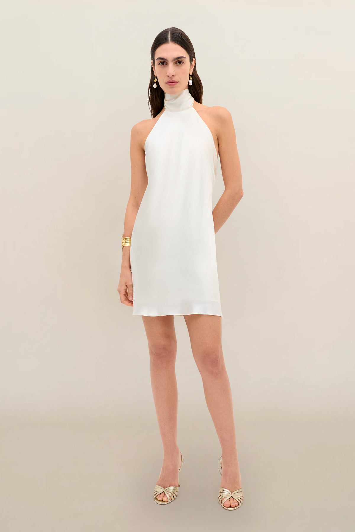 A woman wearing the Capucine - Champagne sleeveless, high-neck, short white silk-satin party dress with an open back, styled with gold earrings, a gold bracelet, and gold open-toe heels, stands against a neutral background.