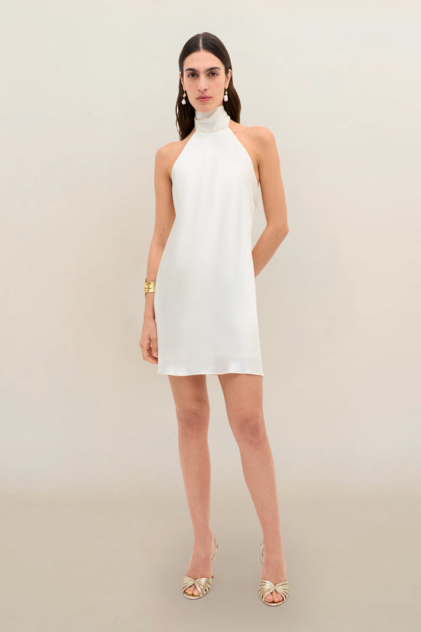 A woman wearing the Capucine - Champagne sleeveless, high-neck, short white silk-satin party dress with an open back, styled with gold earrings, a gold bracelet, and gold open-toe heels, stands against a neutral background.