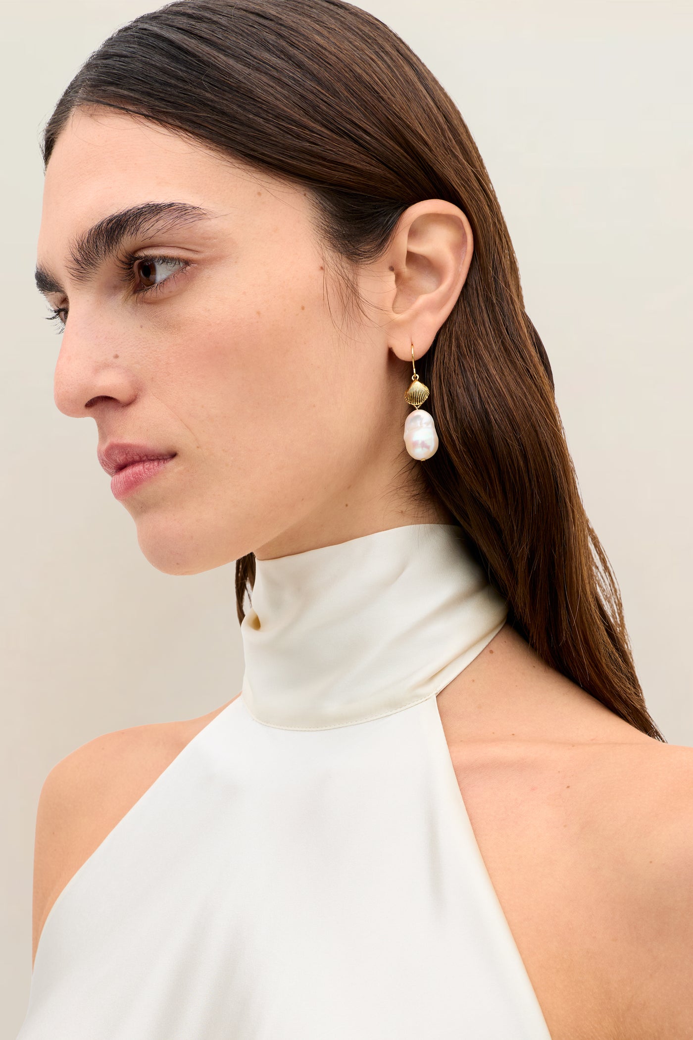 A woman with straight brown hair is shown in profile against a plain, light background, wearing the Capucine - Champagne white silk-satin halter-neck top with an open back and a single gold earring featuring a large pearl.
