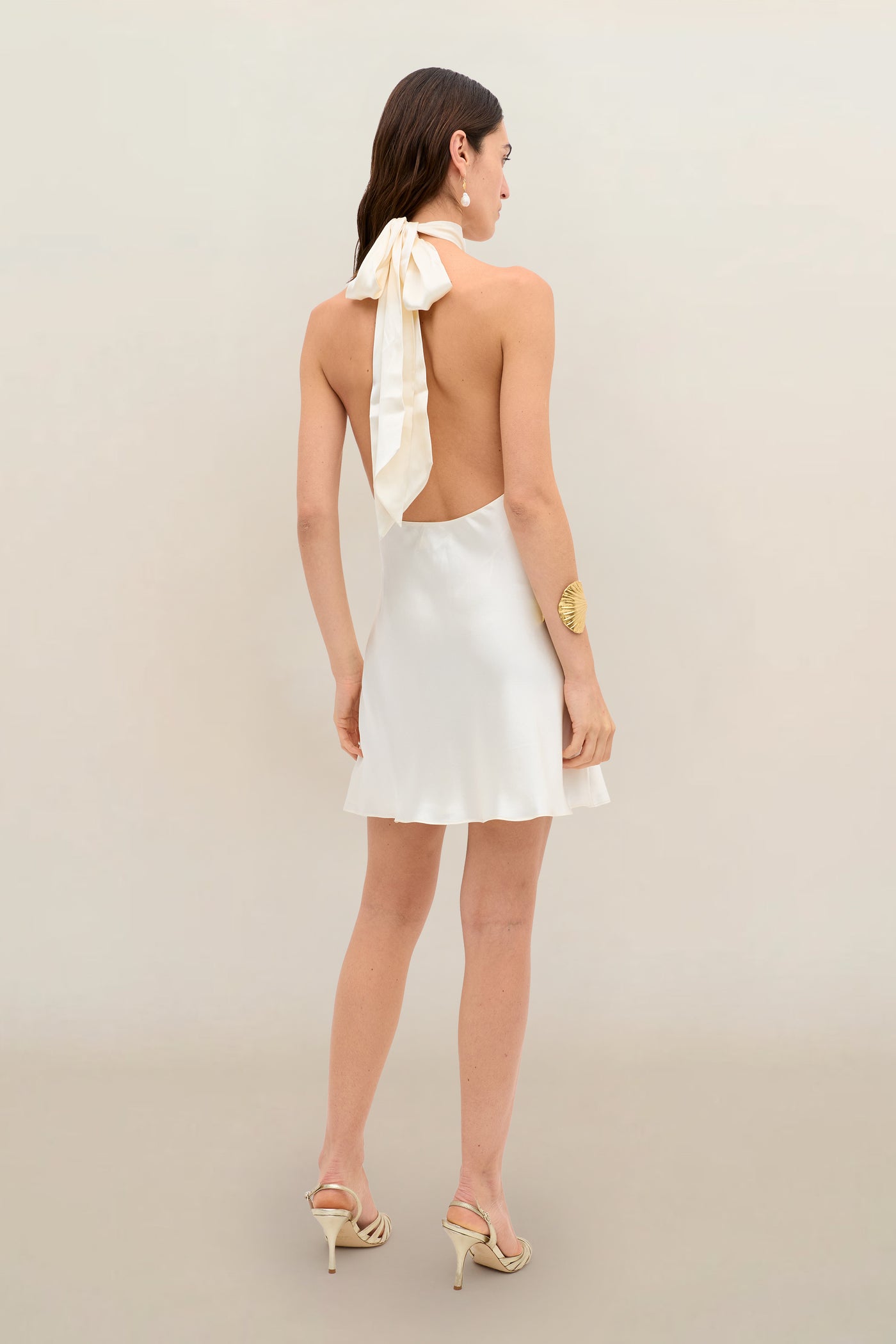 A woman wearing the Capucine - Champagne, a sleeveless white silk-satin halter mini party dress featuring an open back and a large bow tied at the neck, paired with strappy heels, stands against a plain light background.