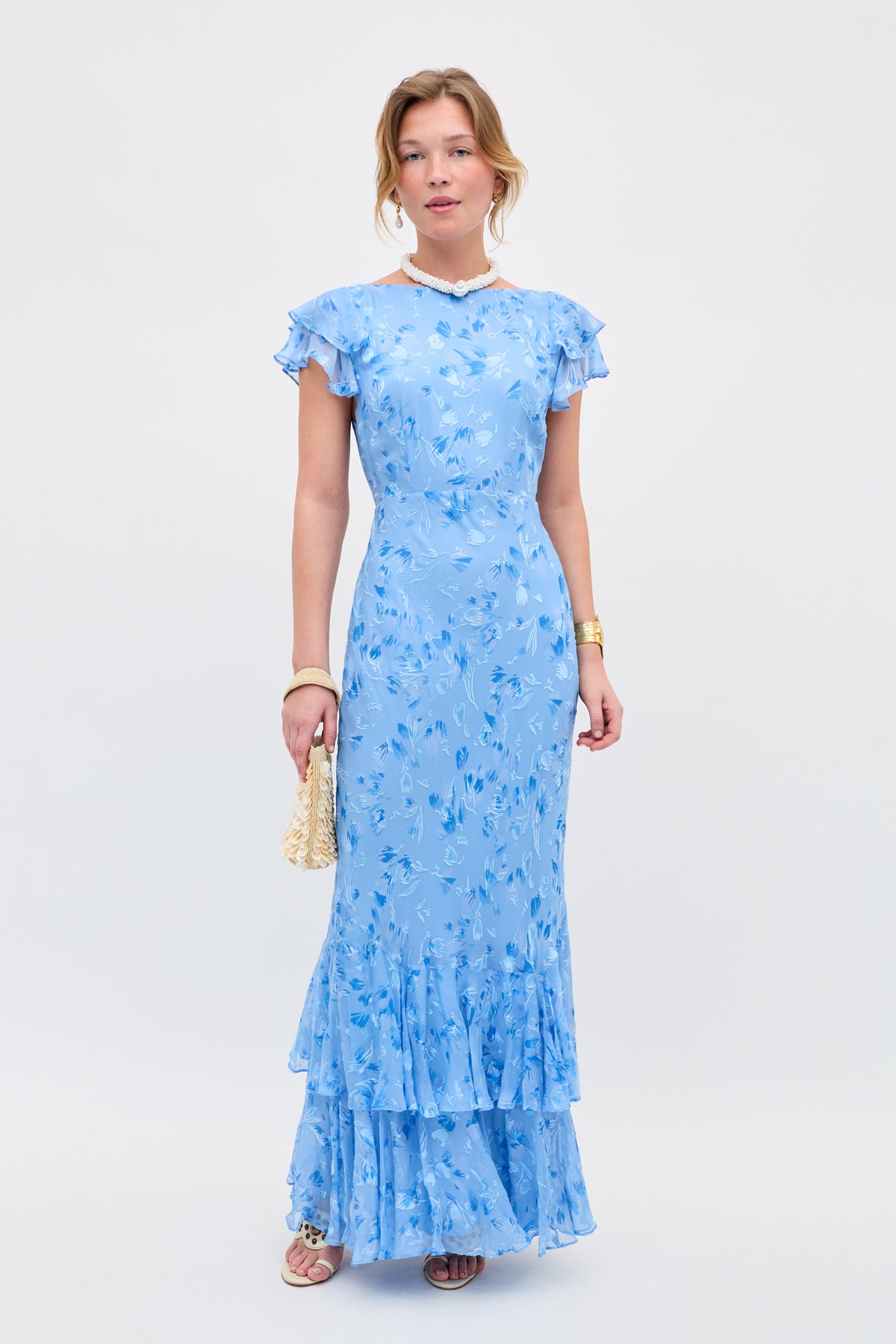 A woman stands against a white background wearing the Ordinia - Ava Devore Blue, a light blue floral maxi dress with ruffled sleeves and hem. She accessorizes with a small woven handbag, sandals, bracelet, and necklace.