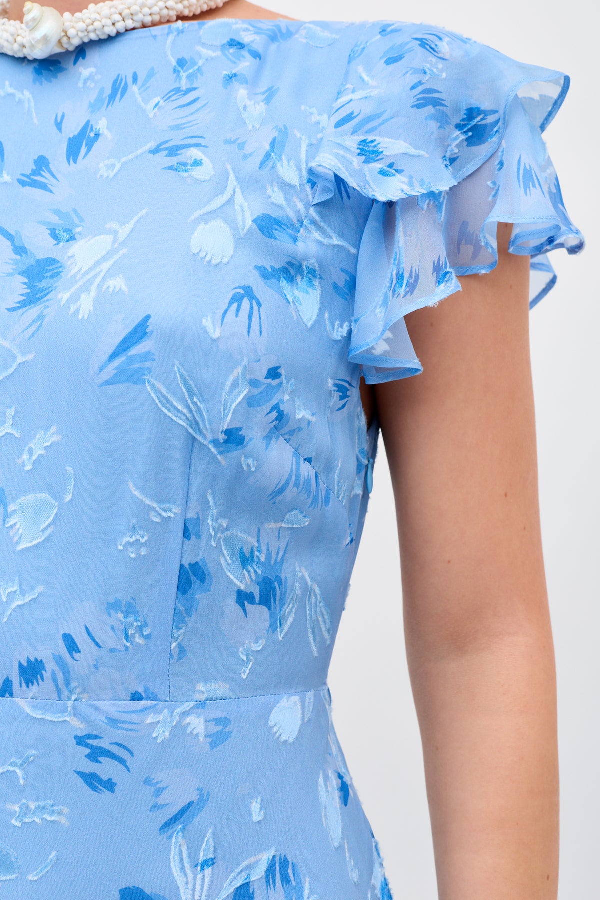 A close-up of a person wearing the Ordinia Ava Devore Blue dress, featuring ruffled short sleeves, a pearl necklace, and a textured floral pattern in shades of blue and white.