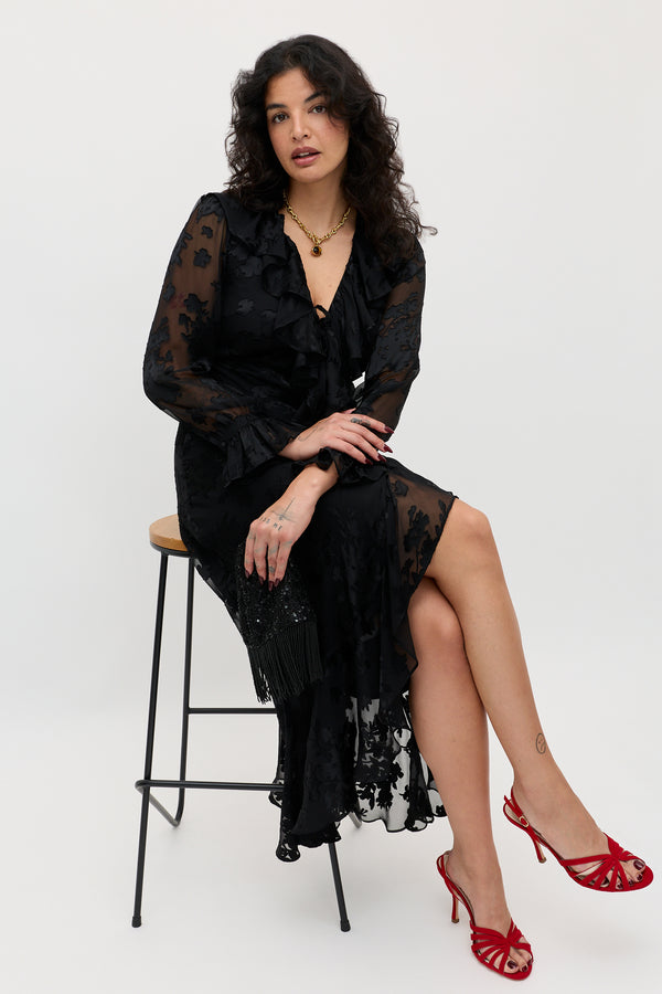 A woman wearing the Gracye – Bouquet Devoré Black, a sheer black dress adorned with floral patterns, sits on a stool with her legs crossed. She accessorizes with red high-heeled sandals and gold jewelry, her wavy dark hair framing a neutral expression against a plain background.