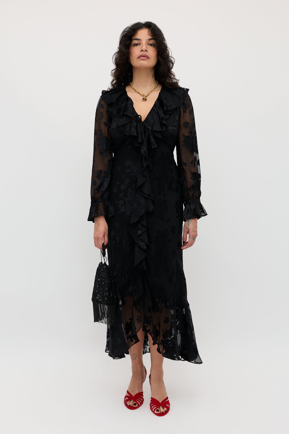 A person wearing the Gracye - Bouquet Devoré Black sheer ruffled dress, red open-toe heels, and holding a black handbag, stands against a plain white background.
