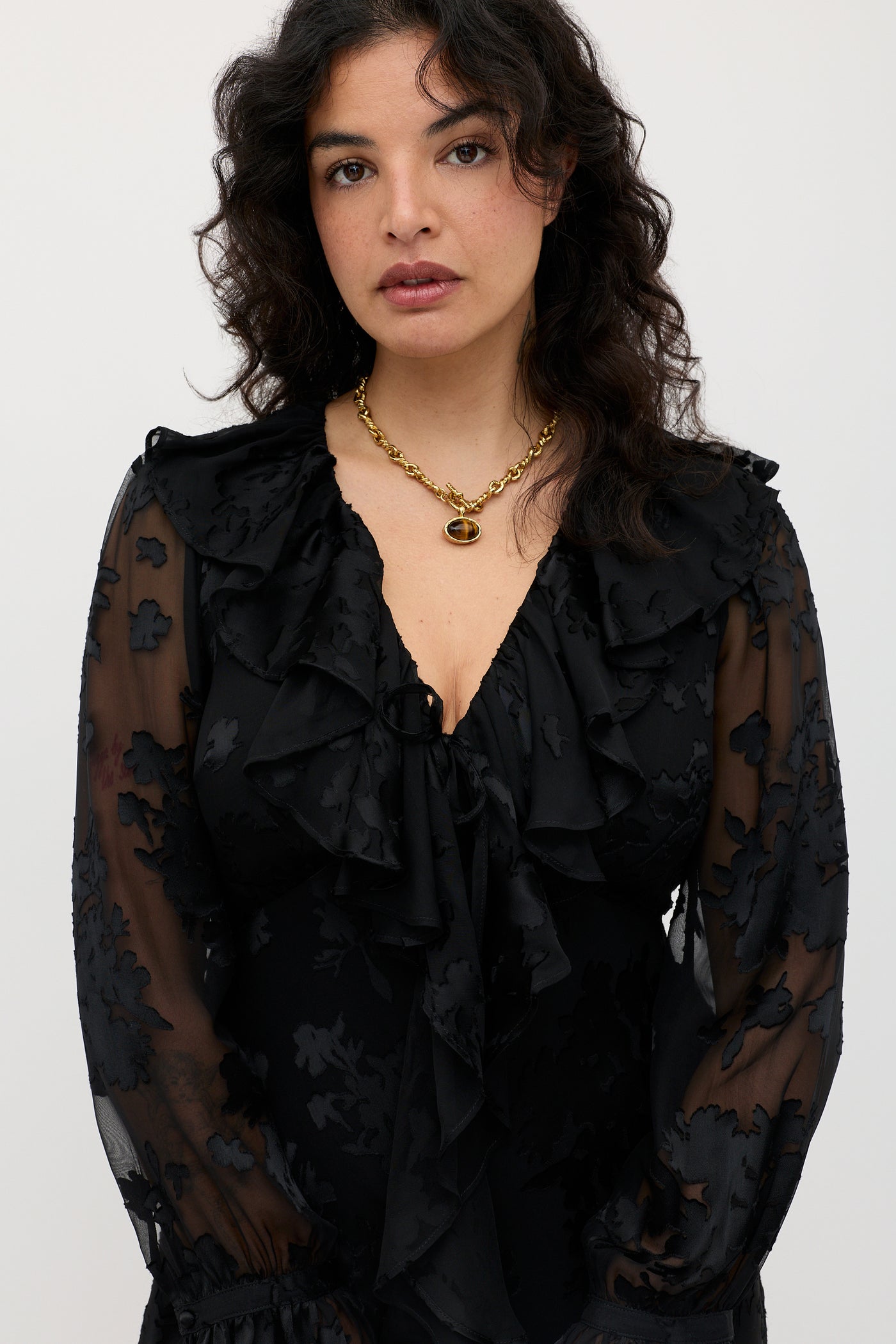 A woman with wavy dark hair wearing the Gracye - Bouquet Devoré Black dress, a sheer black floral design with ruffles, styled with a gold necklace and standing against a plain light background.