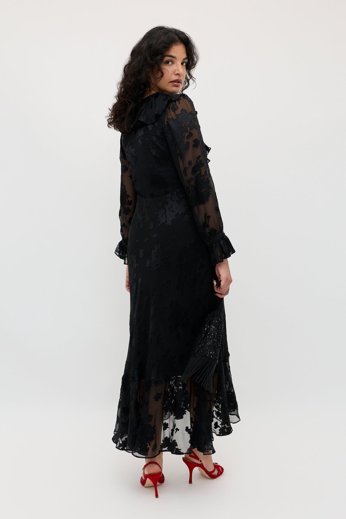 Wearing the Gracye - Bouquet Devoré Black, a woman poses in a long, sheer black floral dress with ruffled cuffs and hem. She stands turned slightly away, glancing over her shoulder in red high-heeled sandals against a plain, light-colored background.