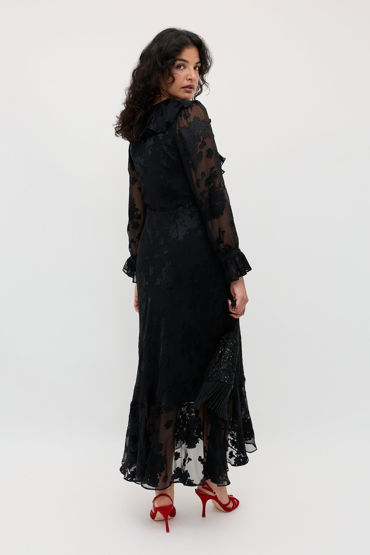 Wearing the Gracye - Bouquet Devoré Black, a woman poses in a long, sheer black floral dress with ruffled cuffs and hem. She stands turned slightly away, glancing over her shoulder in red high-heeled sandals against a plain, light-colored background.