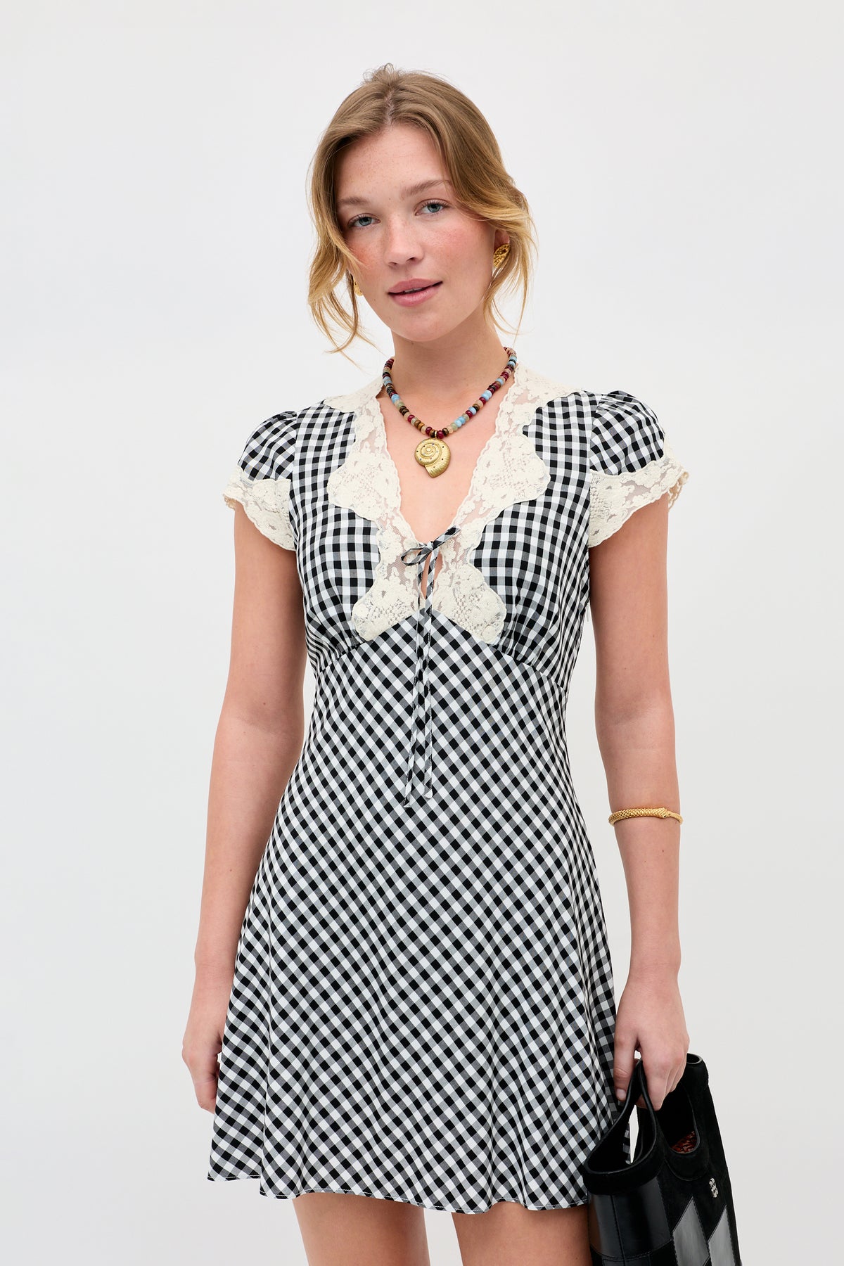 A woman stands against a plain white background wearing the Lacie - Beth Check Black dress with lace trim, paired with a beaded necklace and holding a black handbag.