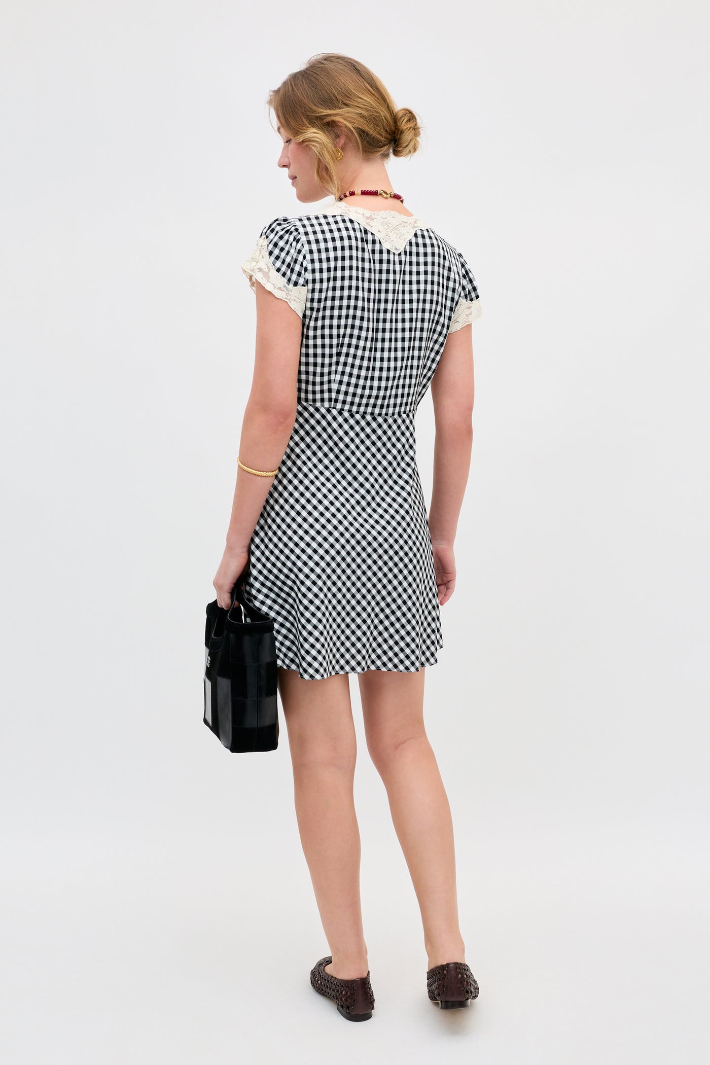 A woman wearing the Lacie - Beth Check Black dress stands facing away, holding a black bag. She has light brown hair in a low bun and wears brown shoes. The background is plain and light-colored.