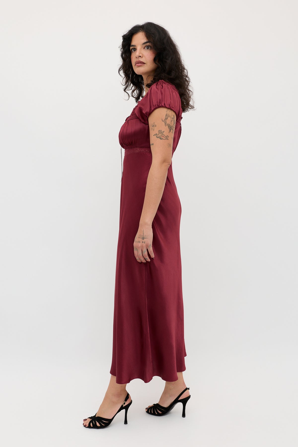 A woman with curly dark hair wears the Maeva - Maroon ankle-length, short-sleeve dress and black heels, standing sideways to show arm tattoos against a plain white background.