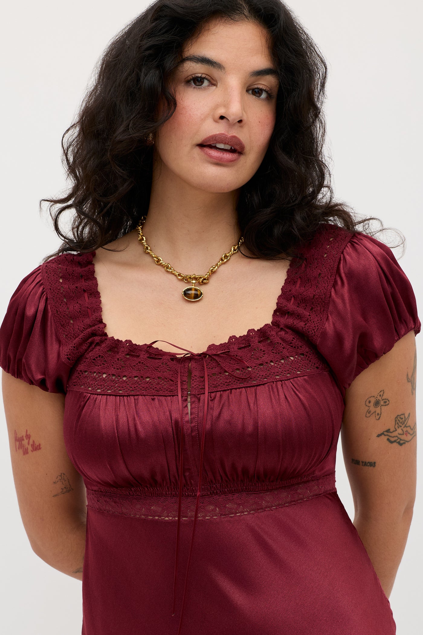 A woman with wavy dark hair wears the Maeva - Maroon dress, featuring puff sleeves and lace details. She accessorizes with a gold chain necklace, shows multiple arm tattoos, and stands against a plain light background.