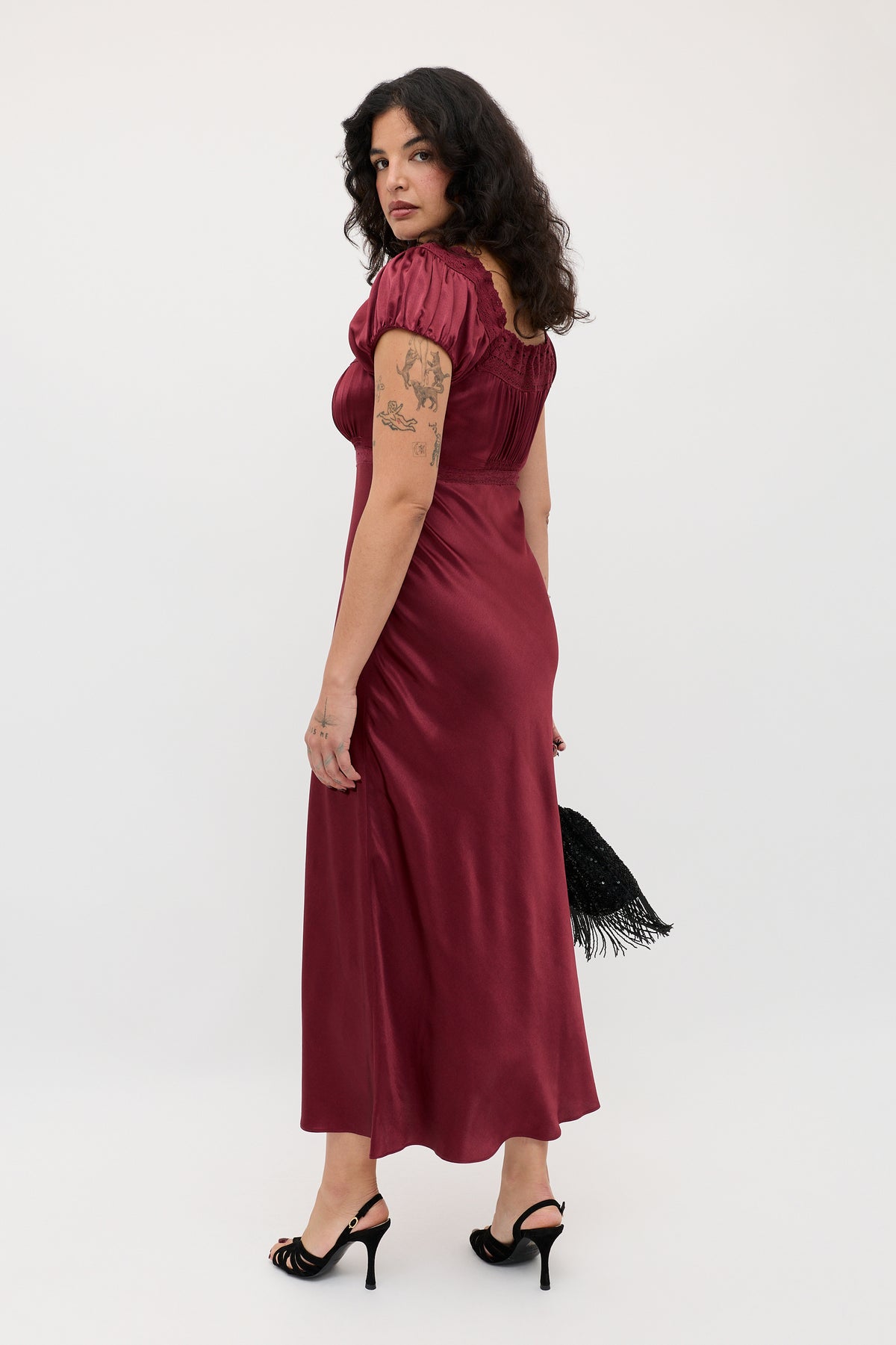 A woman with curly dark hair wears the Maeva - Maroon dress and black heels, holding a black fringed bag. Tattooed on her arm, she stands against a plain white background, glancing over her shoulder.