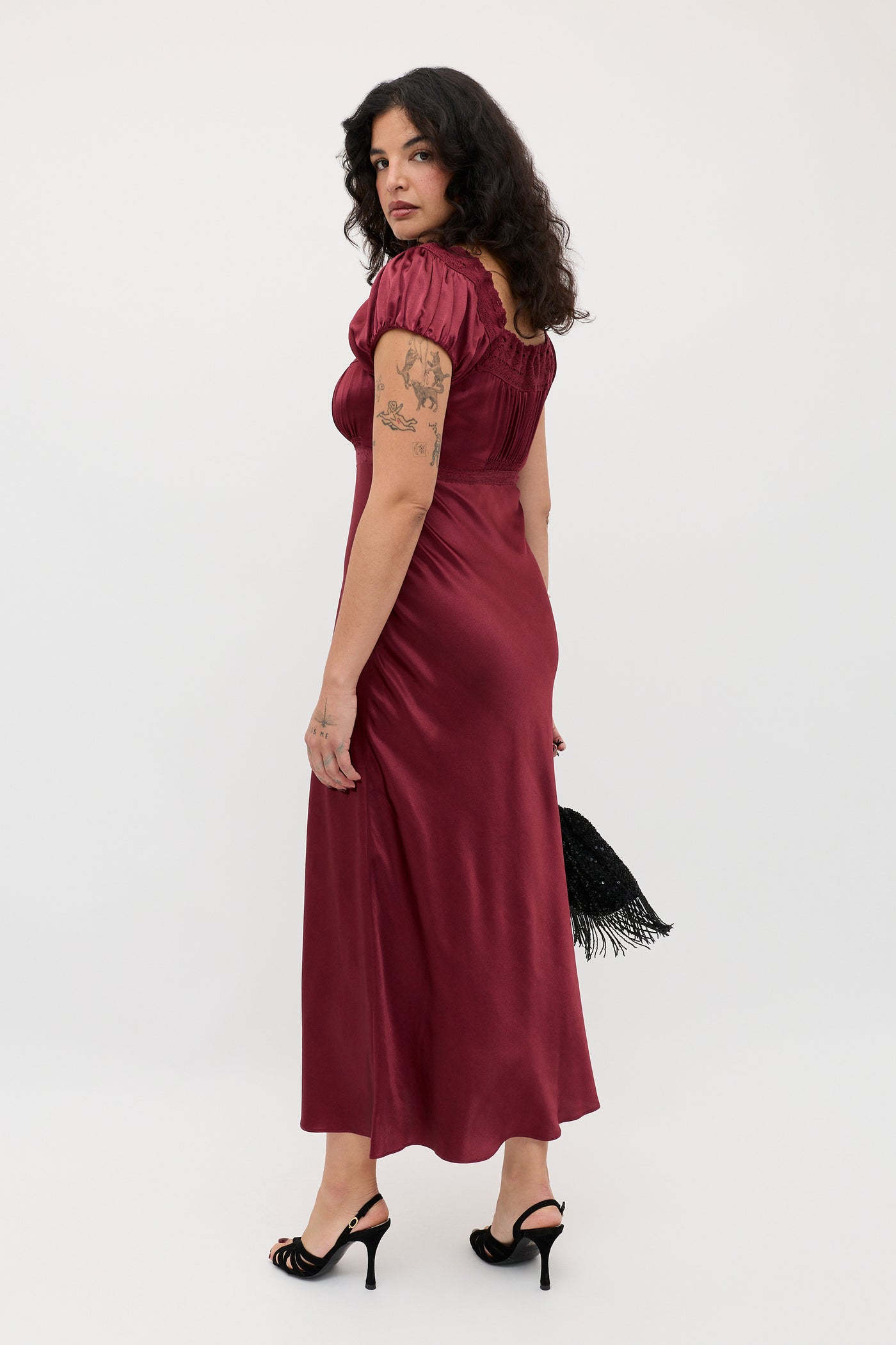 A woman with curly dark hair wears the Maeva - Maroon dress and black heels, holding a black fringed bag. Tattooed on her arm, she stands against a plain white background, glancing over her shoulder.