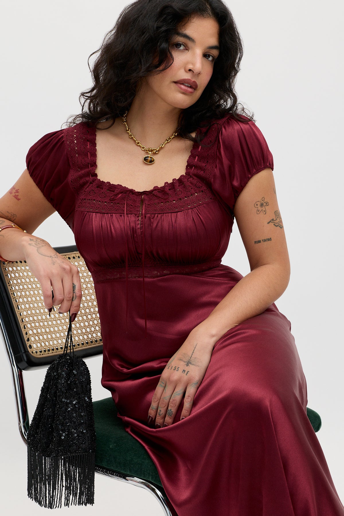Wearing the Maeva - Maroon dress, a woman with wavy dark hair and visible arm tattoos sits on a cane-back chair. She styles it with puff sleeves, a gold necklace, and a black beaded fringe bag.