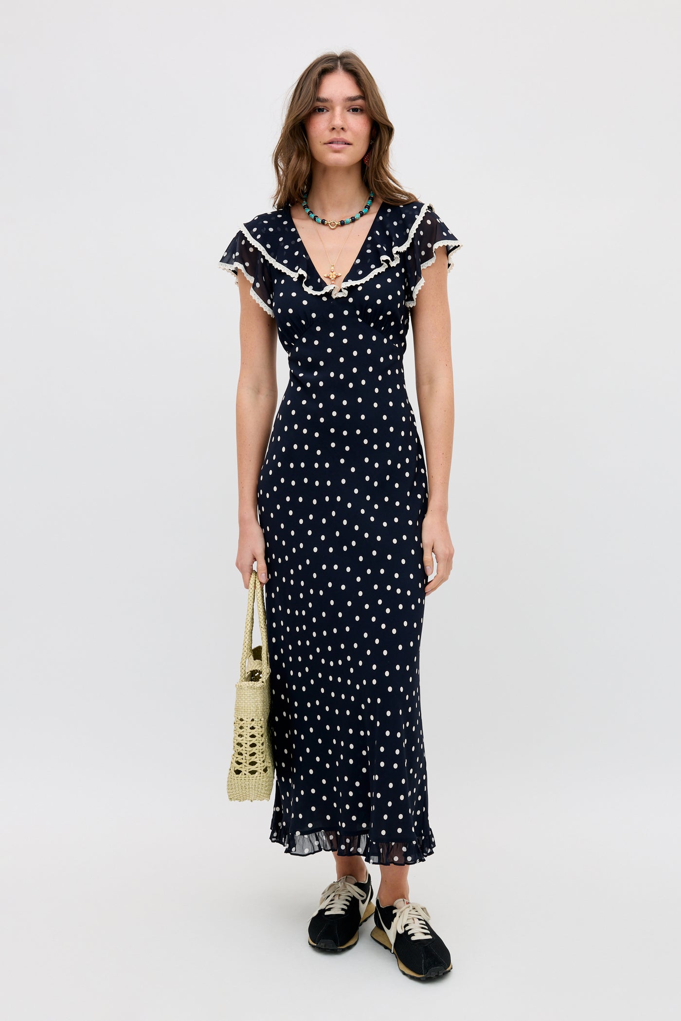A woman in the Bethea - Layla Dot Navy ankle-length polka dot dress with ruffled sleeves stands on a plain light background, holding a woven handbag. She has loose hair, a beaded necklace, and wears black sneakers.