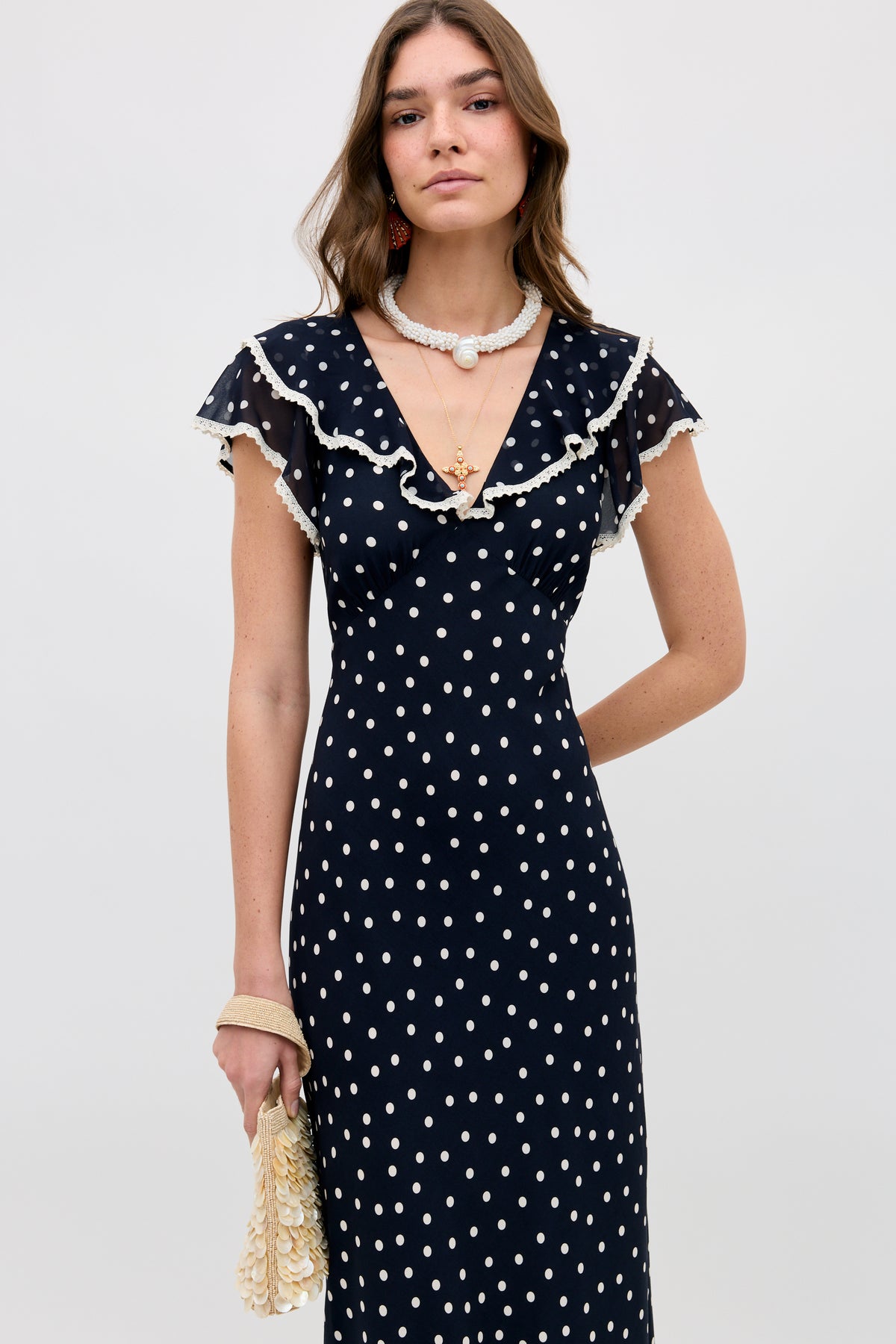 A woman stands against a plain background wearing the Bethea - Layla Dot Navy dress, featuring white polka dots, ruffled short sleeves, and a deep V-neckline. She accessorizes with layered necklaces and a cream-colored handbag.