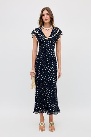 A woman stands against a plain background wearing the Bethea - Layla Dot Navy dress, an ankle-length navy piece with white polka dots, ruffled neckline, and short sleeves. She pairs it with sandals, jewelry, and a small handbag.