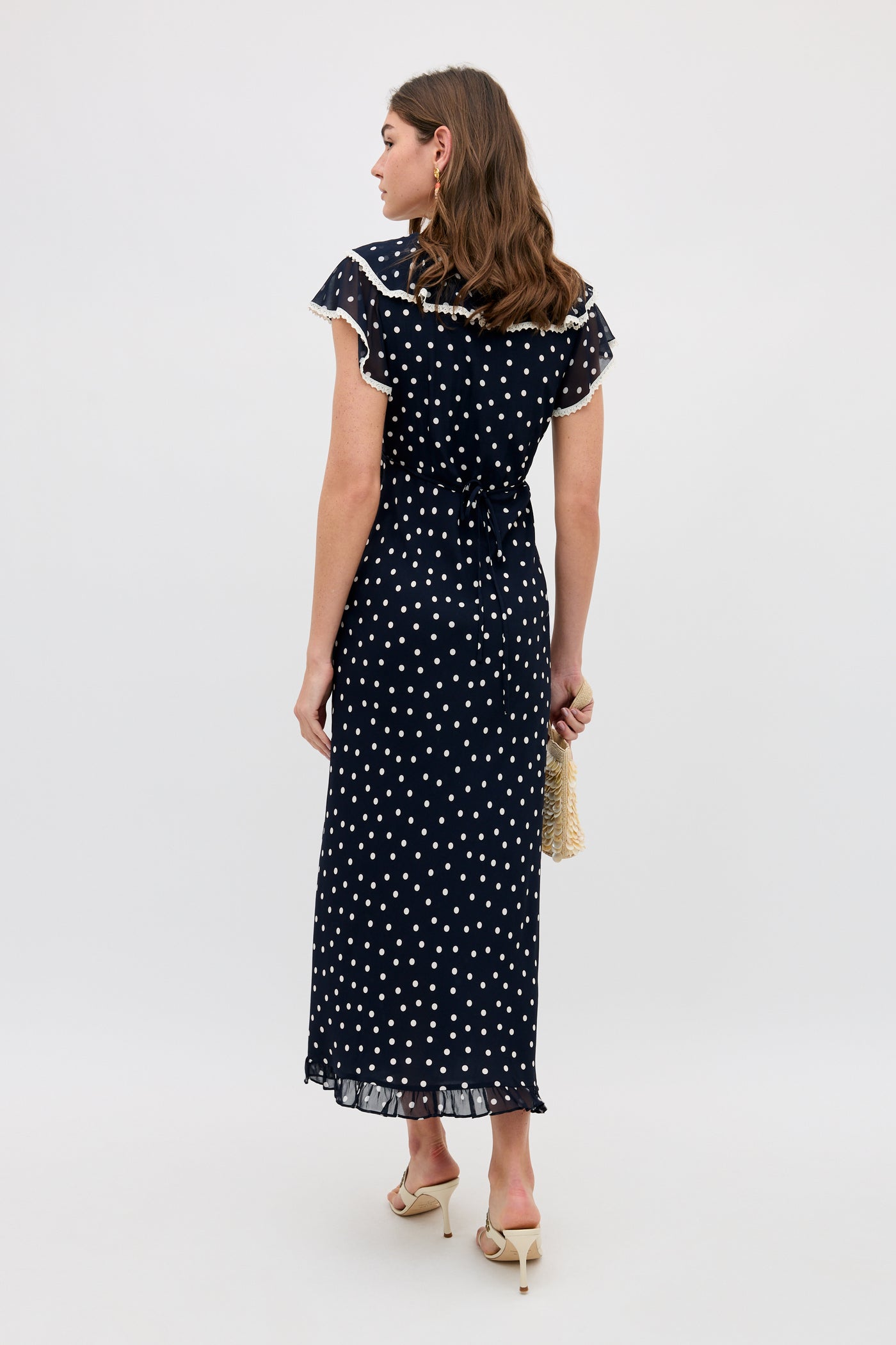 A woman faces away, wearing the Bethea - Layla Dot Navy dress—an ankle-length navy piece with white polka dots and flutter sleeves. She holds a small bag, wears heeled sandals, and stands against a plain light background.