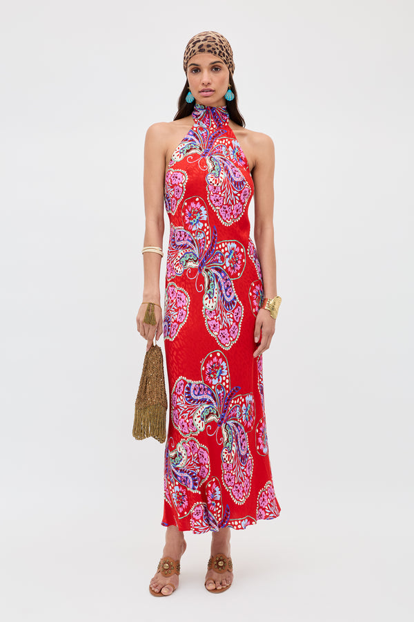 A woman stands facing forward in the Maura - Butterfly Bloom Jacquard Red dress, featuring a vibrant butterfly print. She accessorizes with a leopard-print headscarf, turquoise earrings, gold bracelets, sandals, and a fringed bag.