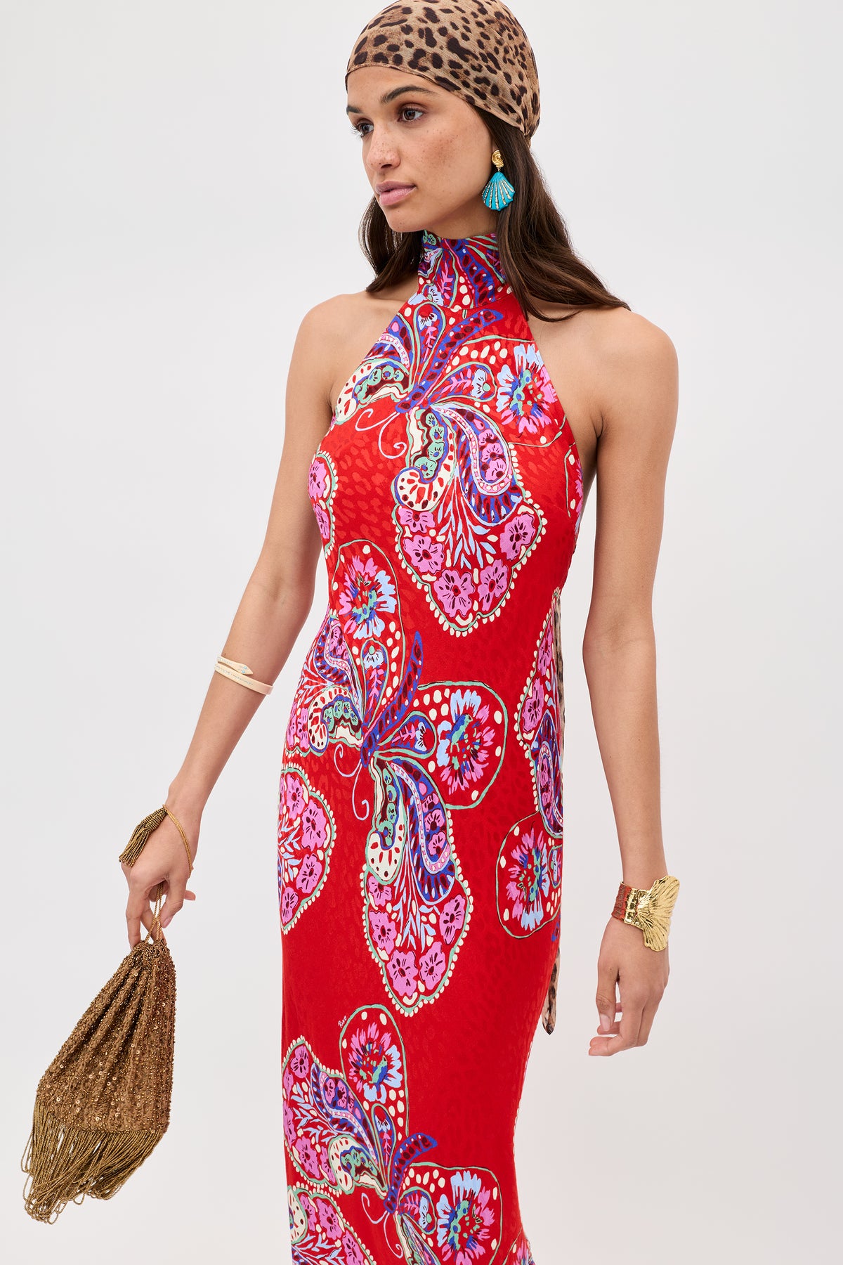 A woman wears the Maura - Butterfly Bloom Jacquard Red dress featuring vibrant florals and butterflies, paired with a leopard-print headscarf, turquoise earrings, gold jewelry, and a woven brown handbag.