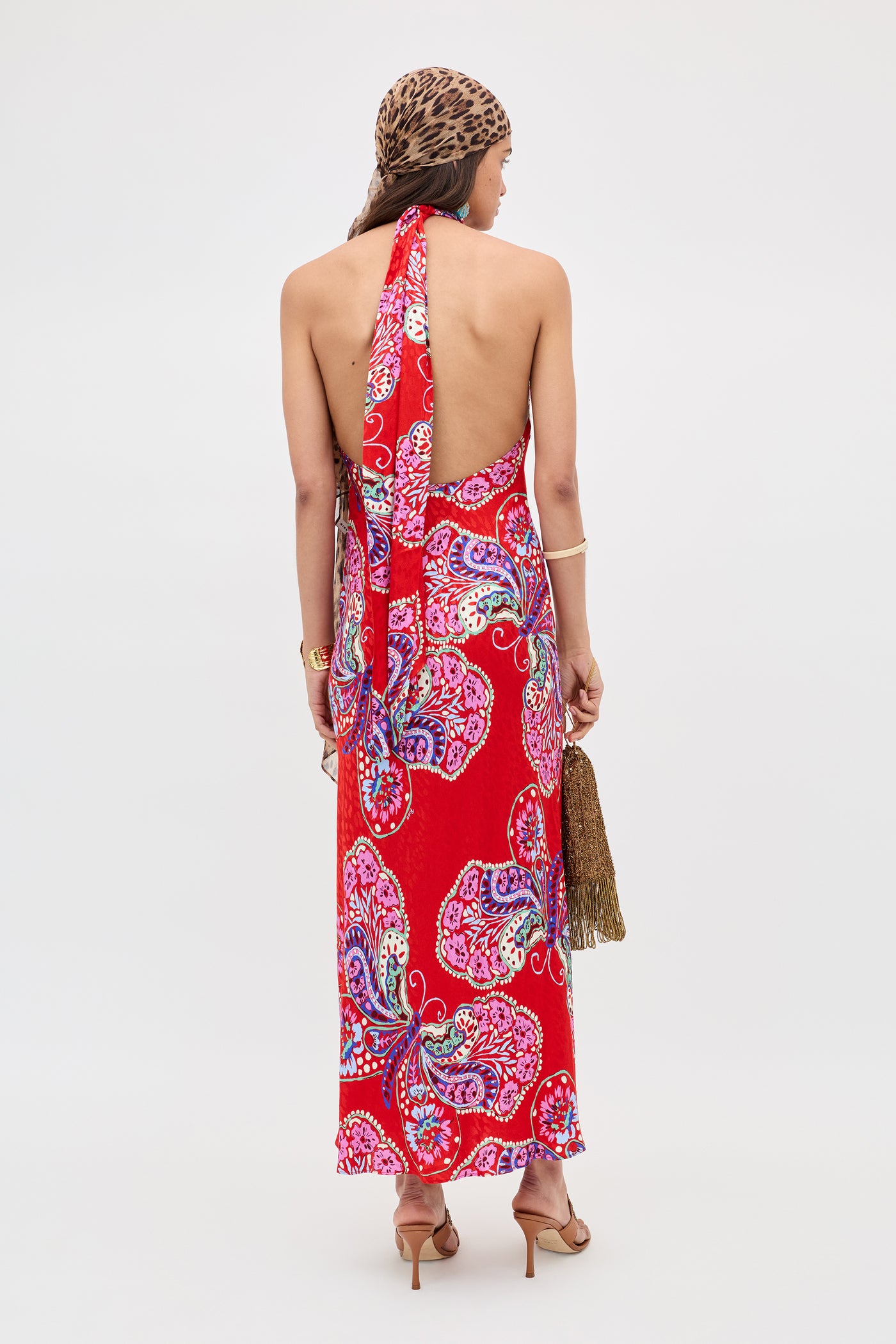A woman faces away in the Maura - Butterfly Bloom Jacquard Red backless halter dress, paired with a leopard-print headscarf, gold jewelry, strappy heels, and holding a brown fringed bag.