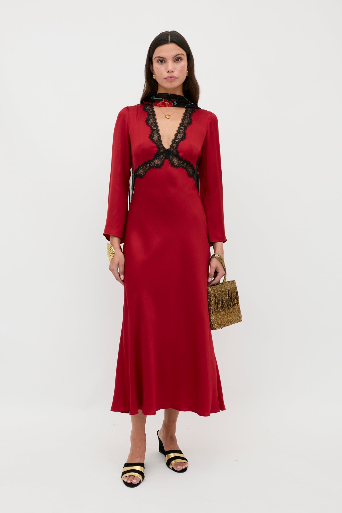 A woman stands against a white background wearing the Debra - Burgundy satin midi dress with black lace trim, paired with black and gold heeled sandals, a gold bracelet, and holding a gold textured handbag.