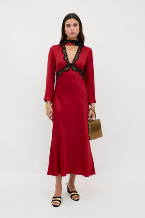 A woman stands against a white background wearing the Debra - Burgundy satin midi dress with black lace trim, paired with black and gold heeled sandals, a gold bracelet, and holding a gold textured handbag.
