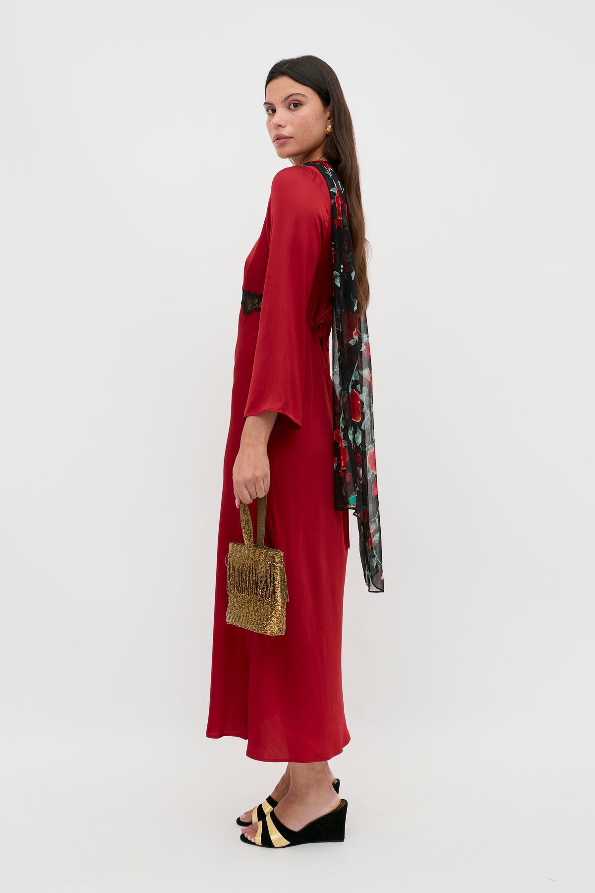 A woman in the Debra - Burgundy dress with wide sleeves stands sideways on a plain white background, holding a woven handbag and wearing black wedge sandals. She has long brown hair and a floral scarf draped over her shoulder.