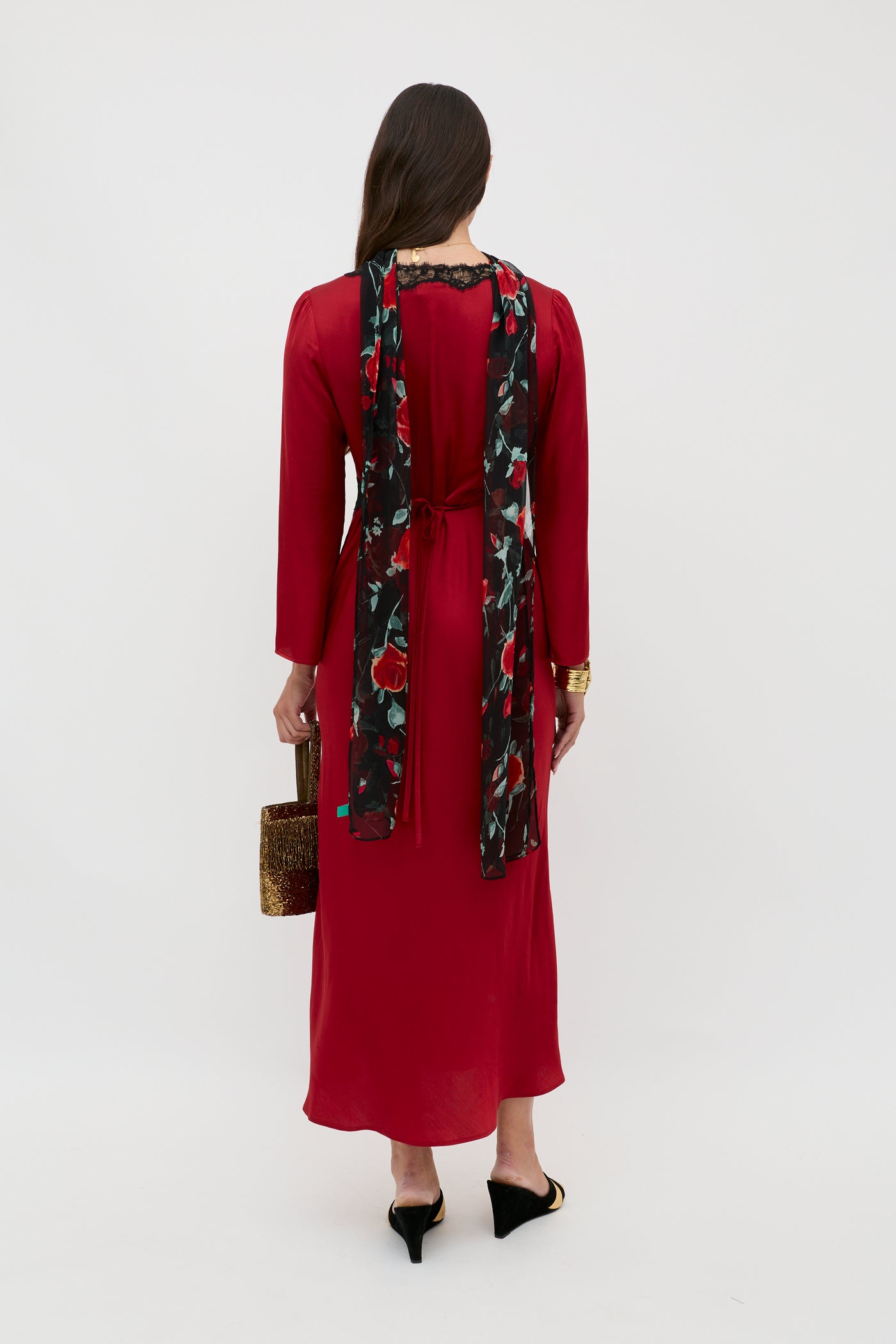 A woman with long brown hair stands facing away, wearing a red dress with a black floral scarf. She carries the Debra - Burgundy textured handbag and completes her look with black heeled mules.