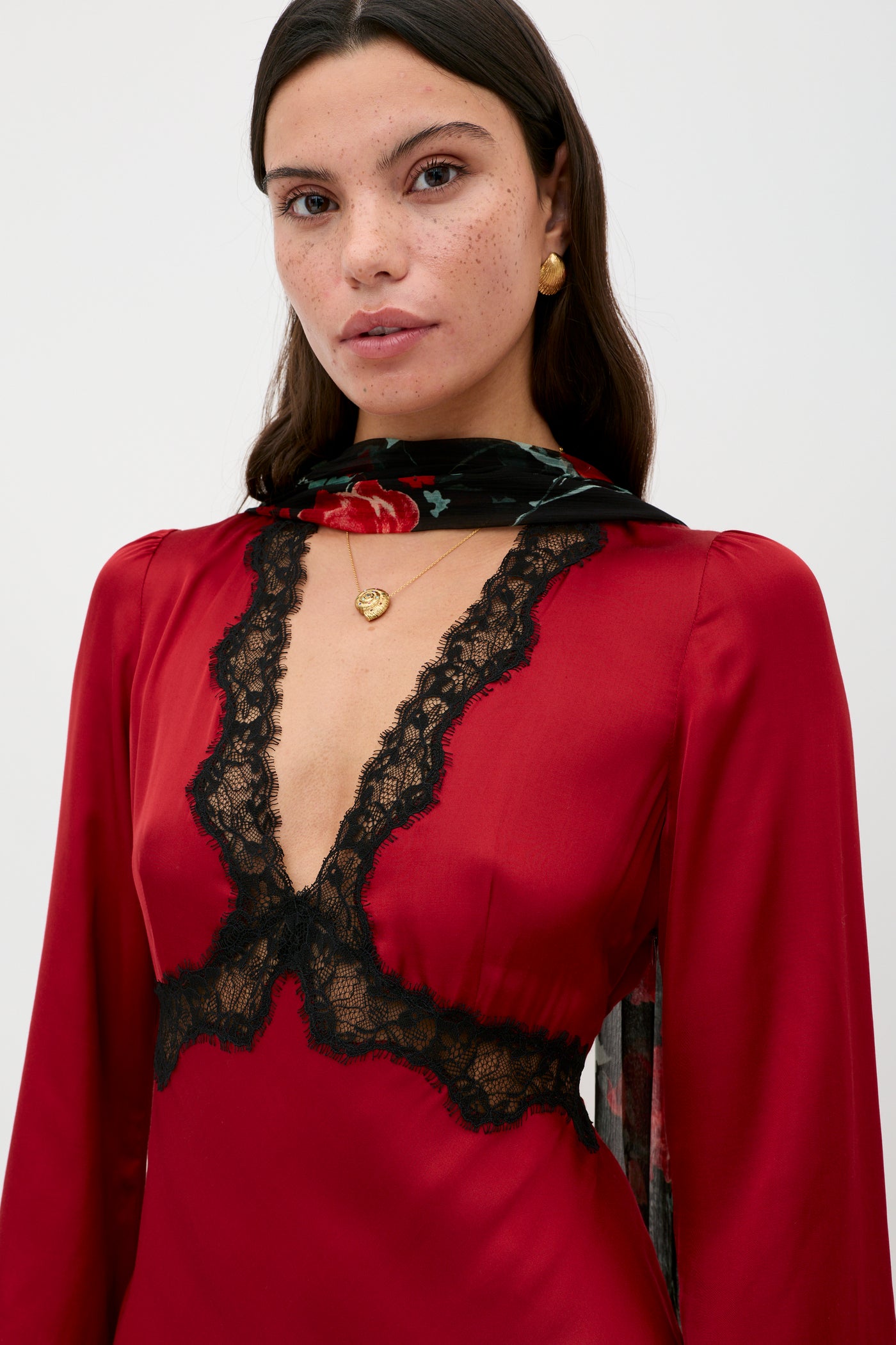 A woman with long dark hair wears the Debra - Burgundy dress, gold earrings, a gold necklace, and a floral scarf. She has freckles and gazes directly at the camera against a plain background.